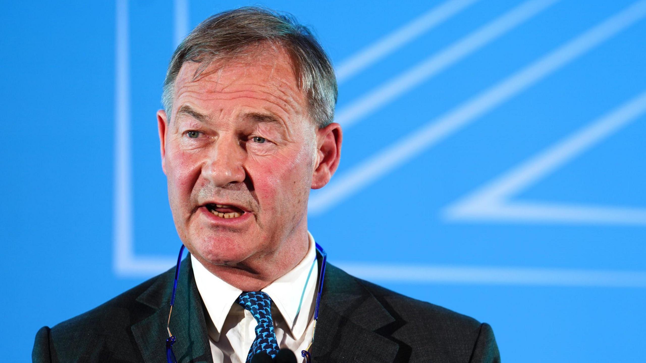 Rupert Lowe is wearing a dark business suit, a white shirt, patterned blue tie and has a pair of glasses hanging round his neck. He is standing at a podium with a microphone. The background is a bright blue presentation screen with abstract white geometric lines.