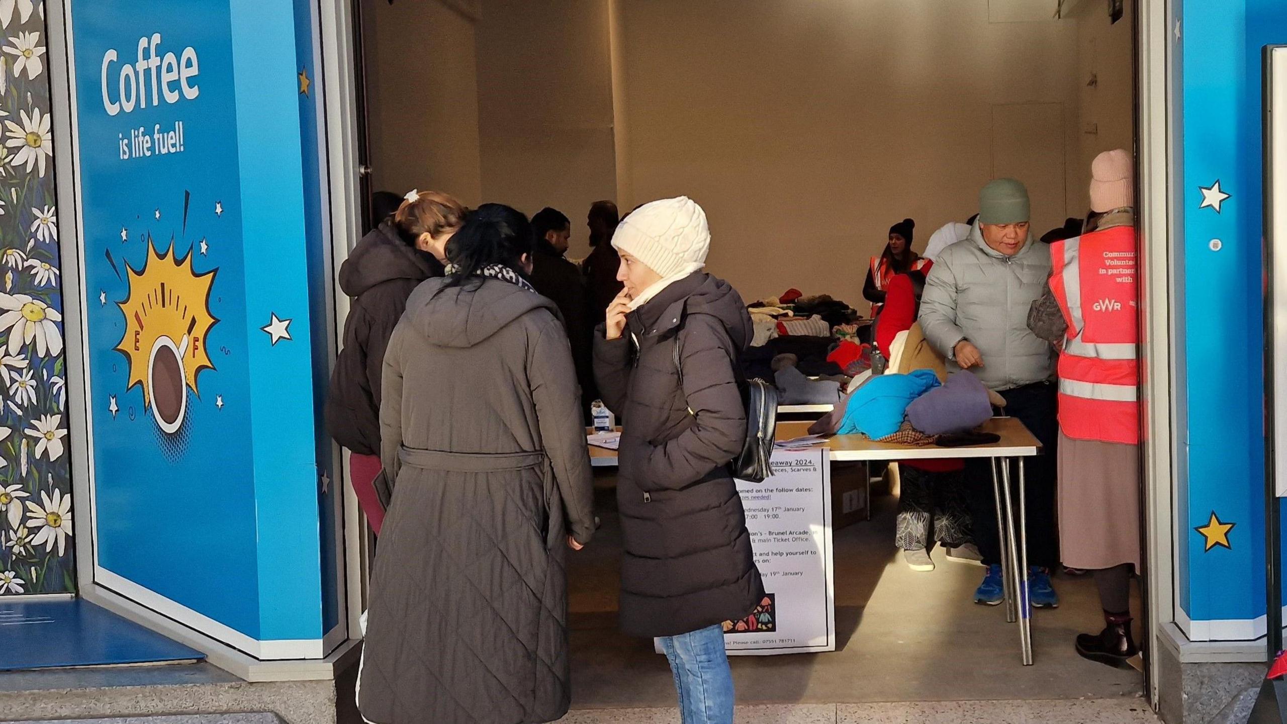 Three women wearing hats and coats standing outside the entrance to the industrial unit having a conversation. Behind them, tables are set up with piles of winter clothes on them, and a woman in a pink high-visibility jacket is standing next to them.