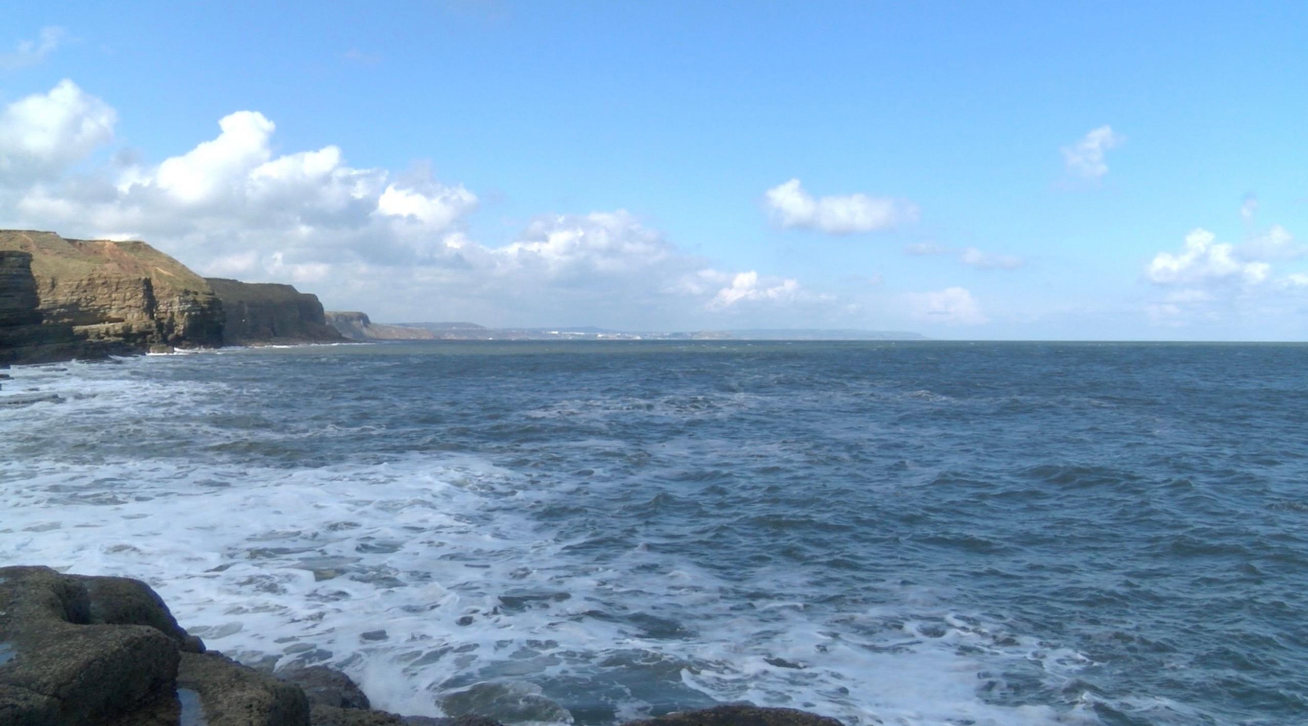 The sea laps rocks on a sunny day in Yorkshire; a grass-topped headland in the distance.