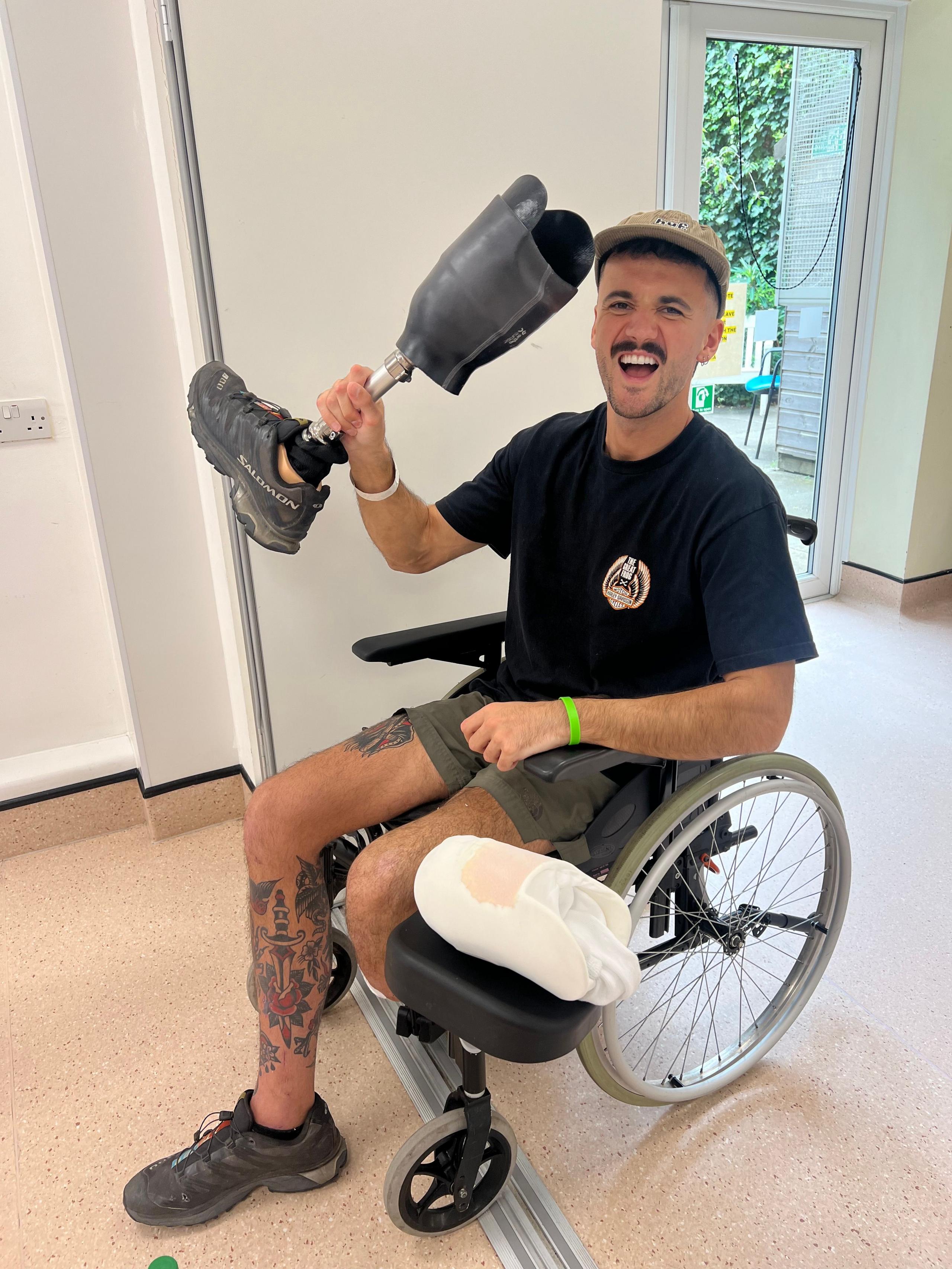 Luke sitting in a wheelchair, holding an artificial leg, he is smiling, with a cap on and looking straight at the camera. He is wearing a black T-shirt, green shorts and you can see one leg with tattoos on and another that has been amputated below the knee. He has padding on the side of the chair. A door is behind him. 