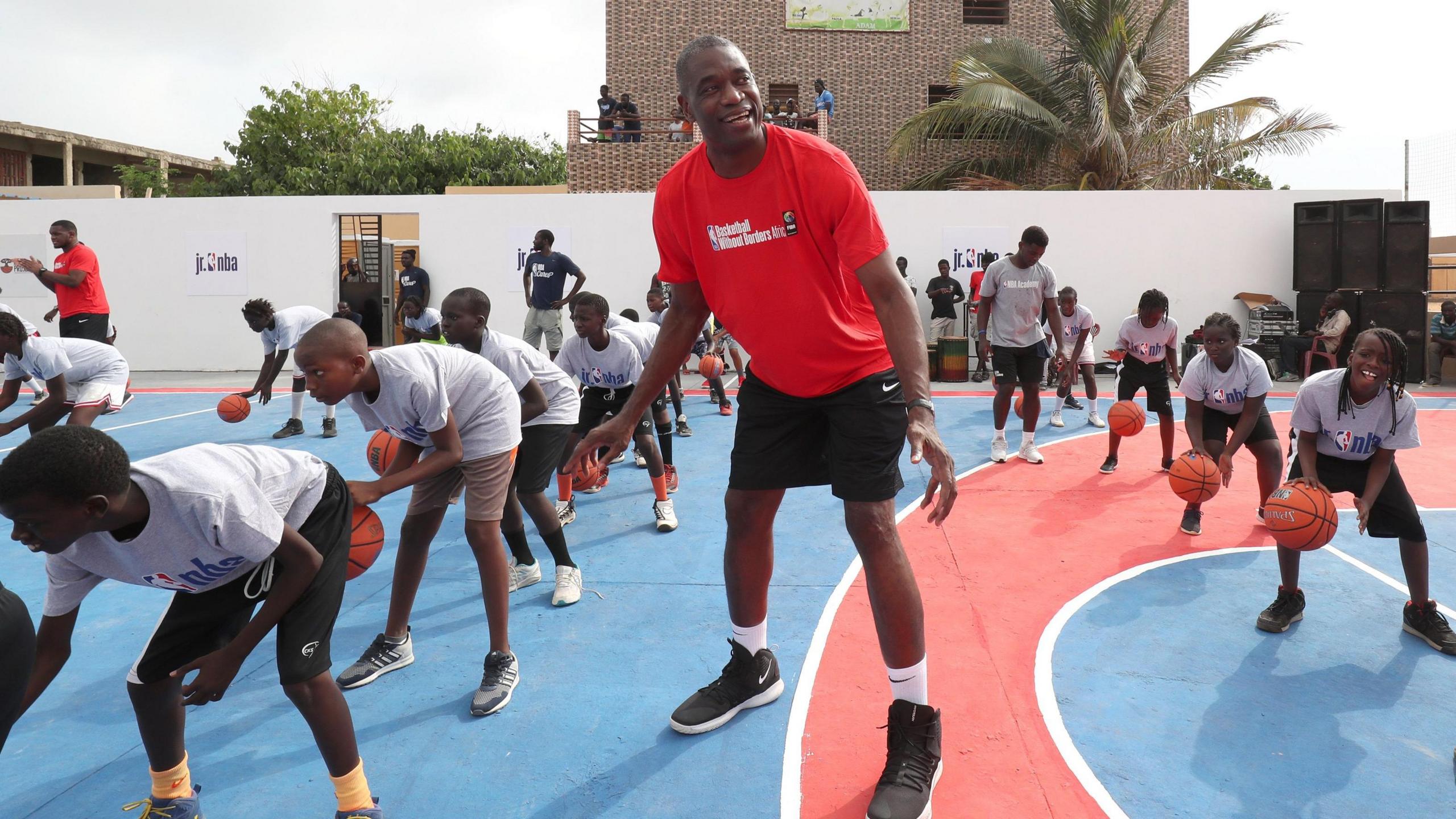 Dikembe Mutombo participates in Basketball Without Borders Africa training camp in July 2019, with several children bouncing basketballs in the background