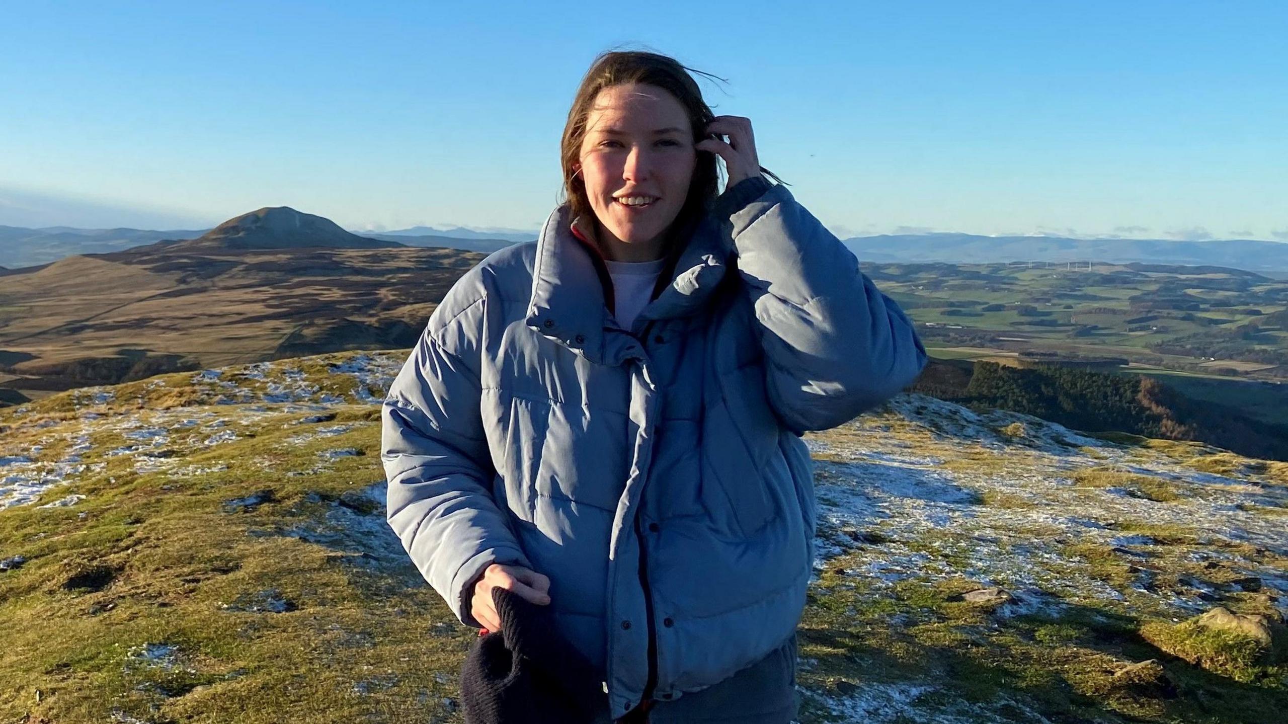 Susie McAdam standing amidst a landscape with fields and mountains in the background, wearing a coat, smiling.