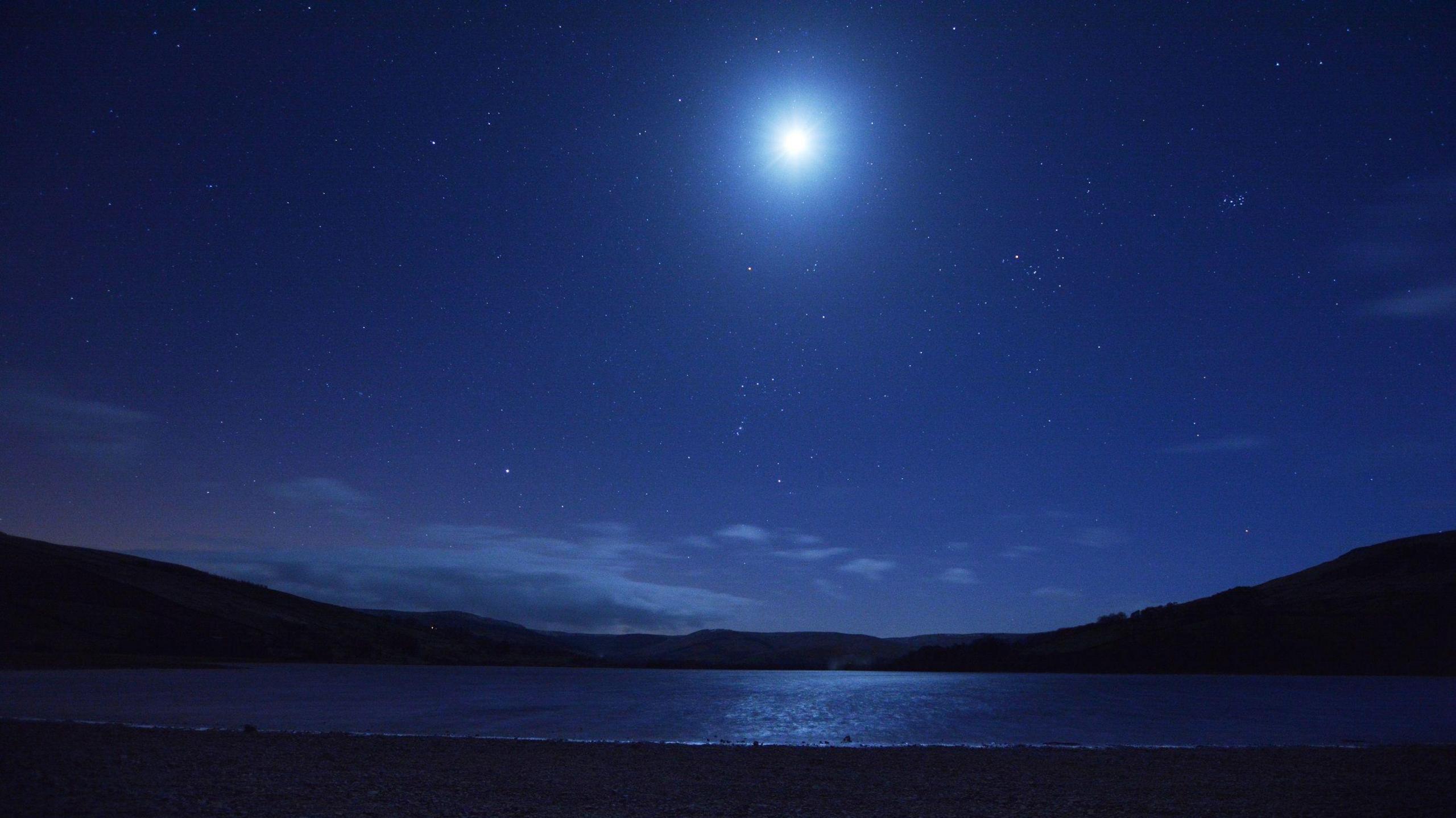 The moon shining brightly in the dark, streetlight-free sky over Semerwater, Wensleydale, in the Yorkshire Dales National Park. The sky appears dark blue. Some low cloud is hanging over the hills which are framing the lake.