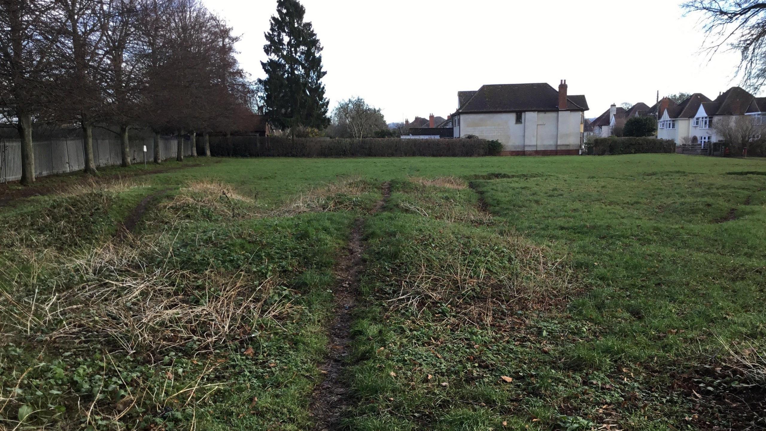 A bike pump track in a residential area. It looks like a green field which has raised mounds with muddy tracks going over it, where people have been riding their bikes. Houses can be seen on a nearby street on the right of the image, and, to the left of the field, there is a fence and a line of trees.