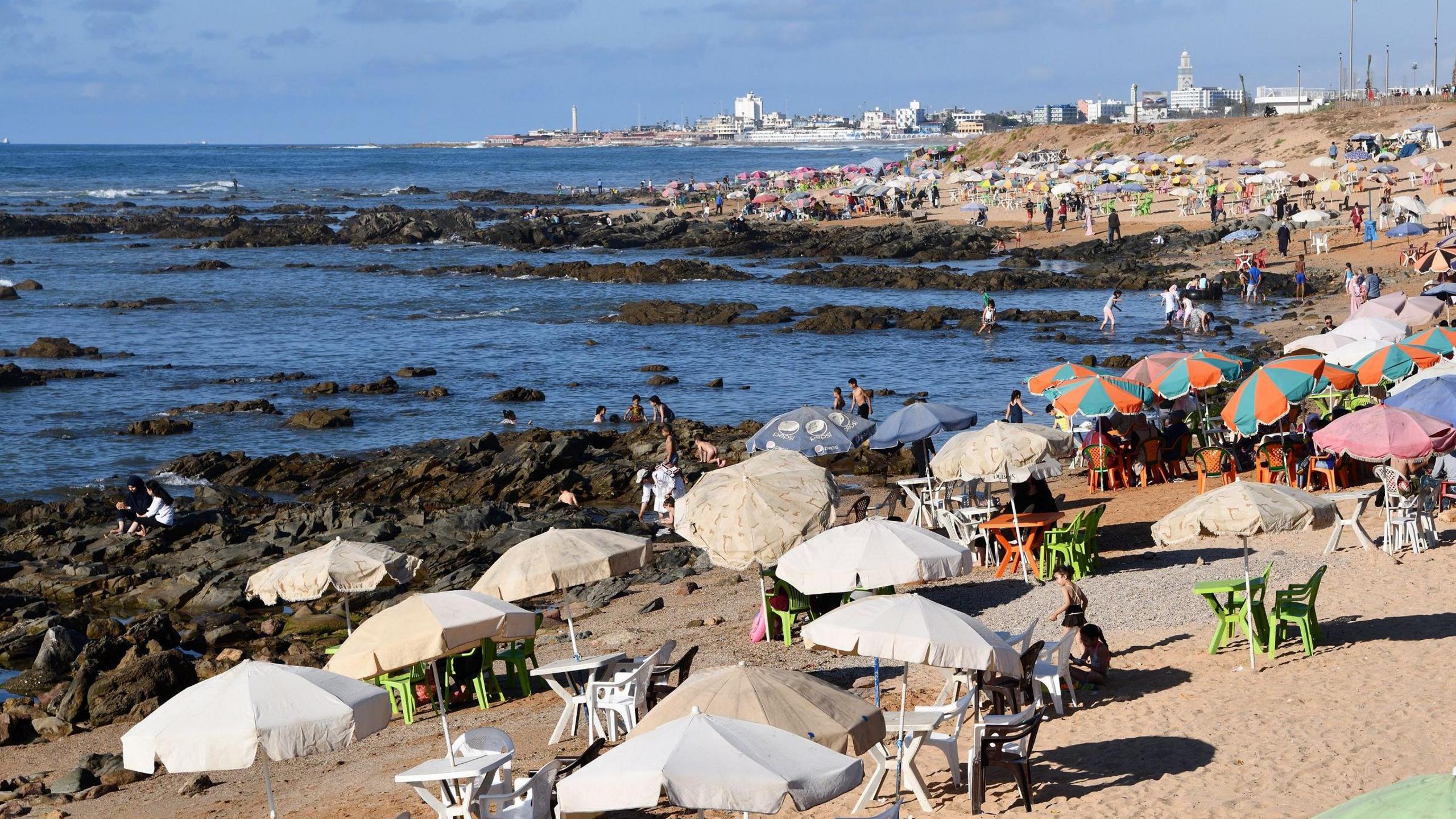 Stock image of people sitting under parasol canopies on the sandy beach behing large rocks next to the sea. Casablanca's buildings appear in the backdrop.