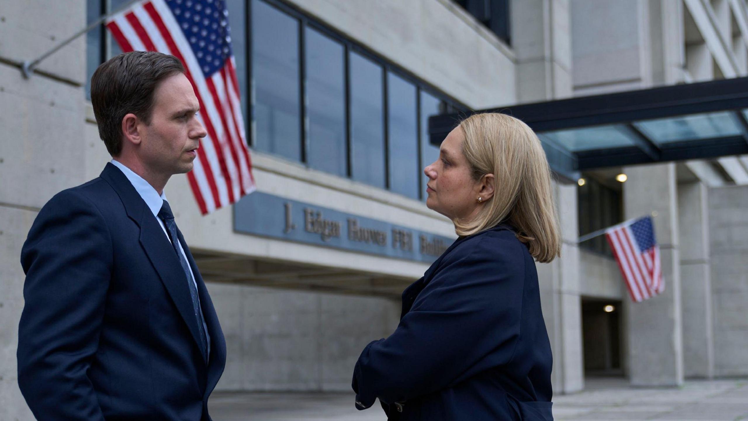 Two actors in dark suits standing outside a US building with stars and stripes flags flying