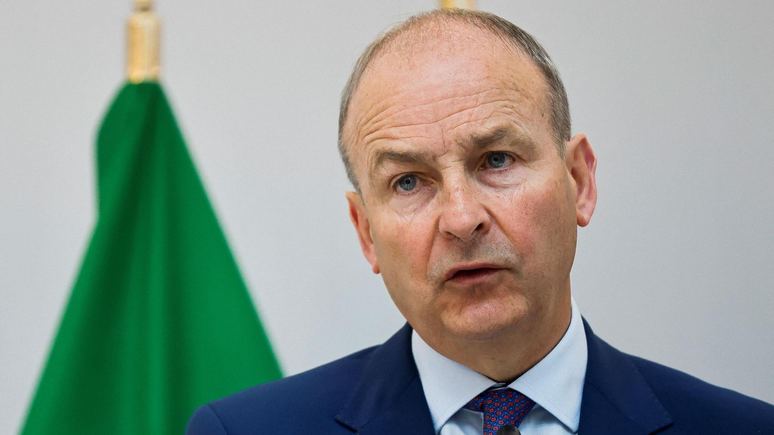 Micheál Martin in navy suit, pictured in front of a green flag