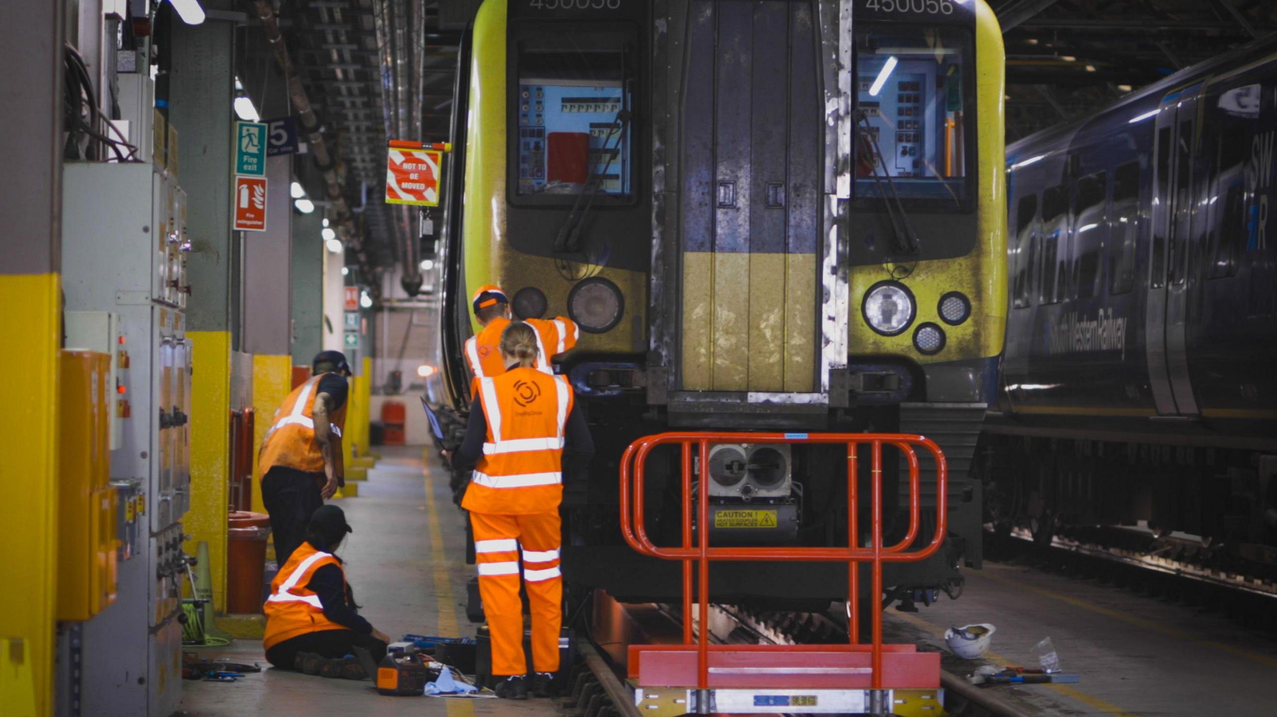 Four railway engineers wearing orange high-vis suits working on the front right of a train in a depot