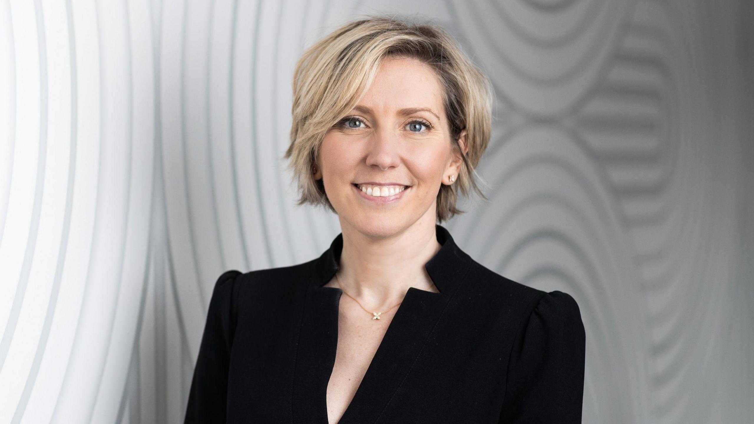 Lindsay Wood is smiling at the camera standing in front of a pale grey background. She has short blonde hair and blue eyes and is wearing a black dress.