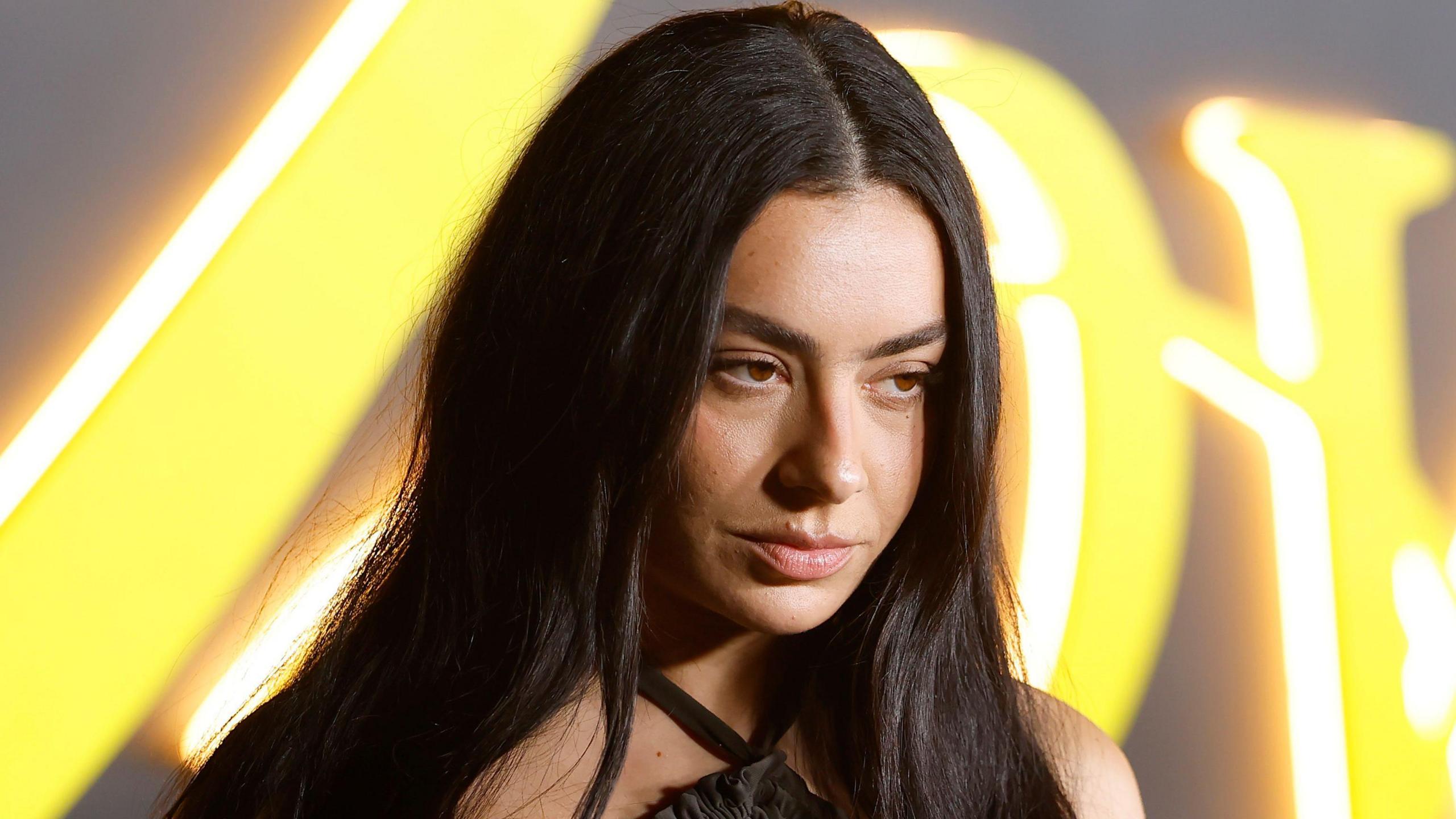 Charli XCX looks just off camera with a serious expression. She has long dark hair down past her shoulders and behind are illegible large letters in neon yellow lights.