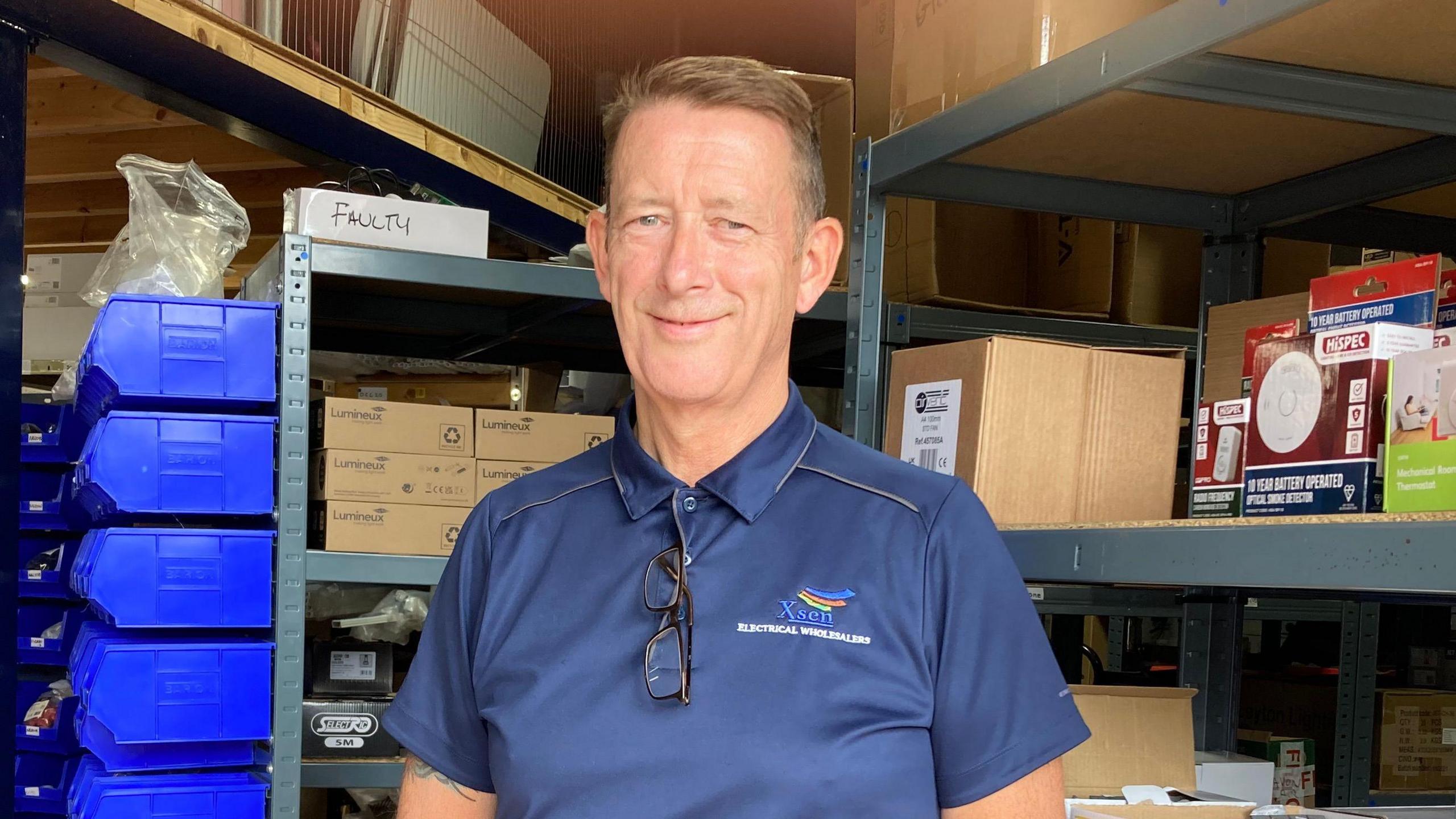 Geoff Spence is stood in the warehouse of his electrical wholesale business. He is wearing a navy blue T-shirt with his company logo on the right-hand side of the photo. His glasses hang from a button hole on his T-shirt. He has short red hair and is smiling at the camera.