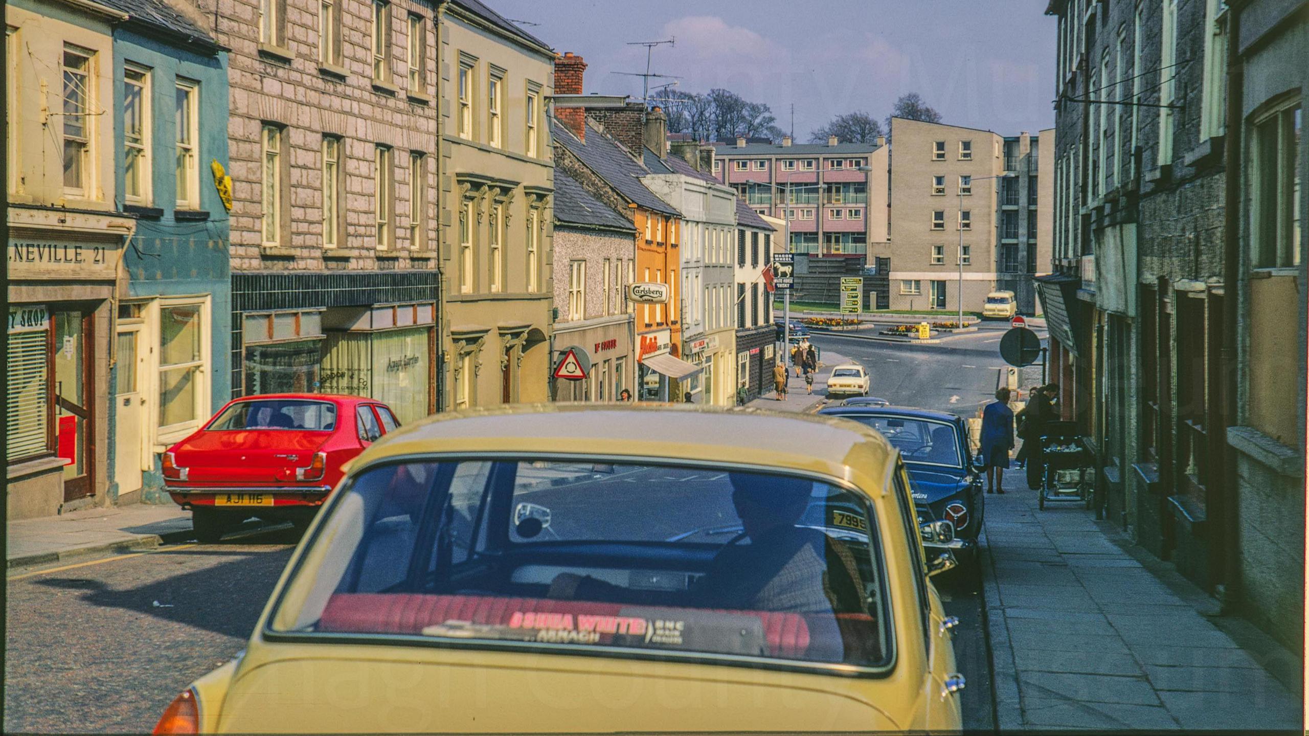 A digitalised photo from a film of a shopping street. The shop fronts are brightly coloured and a yellow car and red car are parked along the street.  Looking east showing the shops at the bottom of Scotch Street and the new development of Bridge House in the background