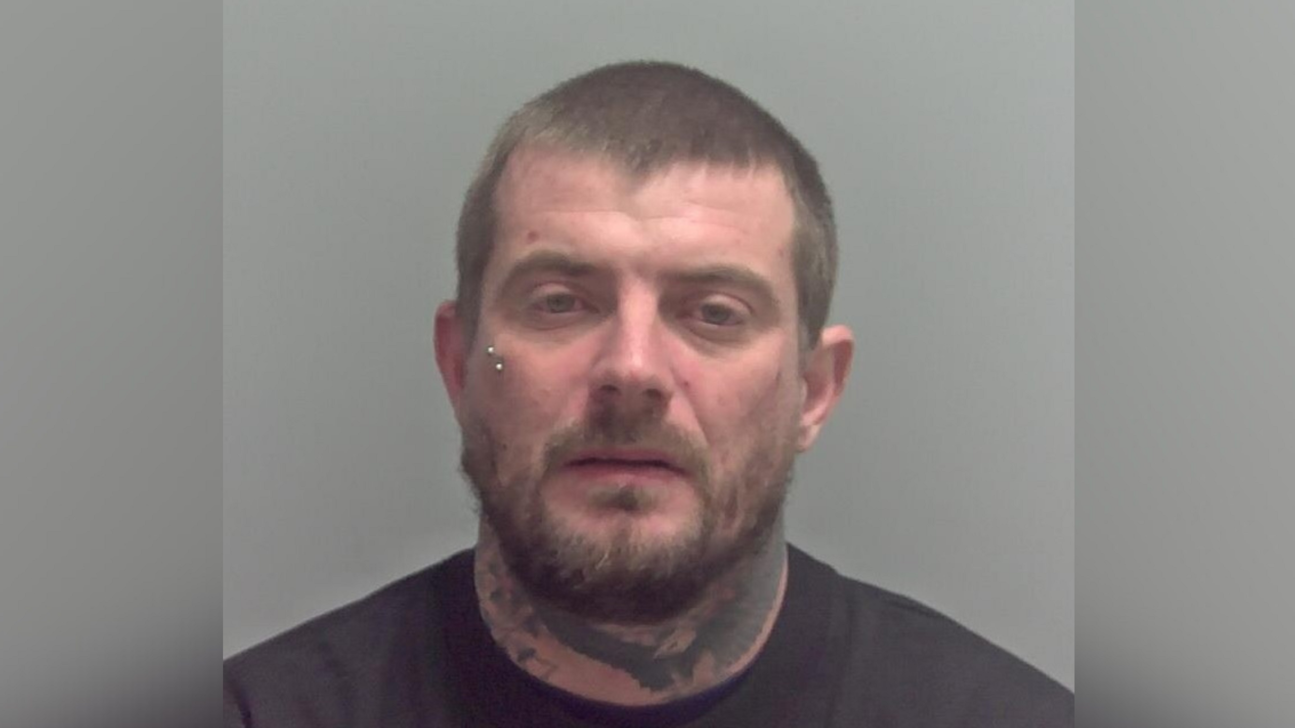 A mugshot image of Ryan Leggett. He is wearing a black T-shirt and has tattoos on his neck. He has a piercing under his right eye. He is looking into the camera.