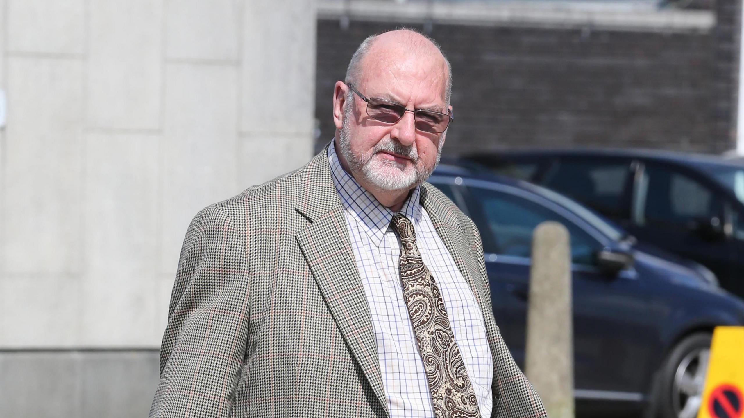 Ifor Whittaker looks at the camera, he is wearing a checked shirt and a paisley tie with a tweed-style jacket. He is wearing glasses and has a short white beard