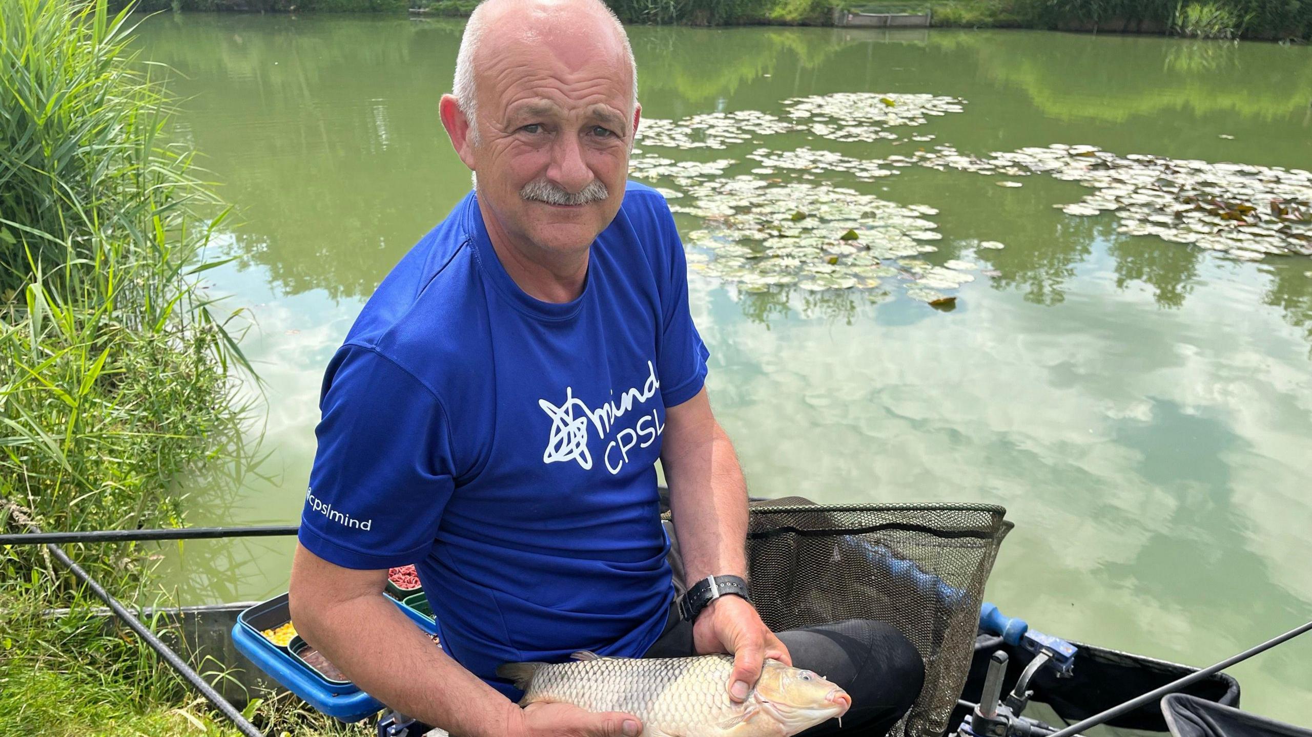 Ian Darler holding a fish while sitting by a lake 