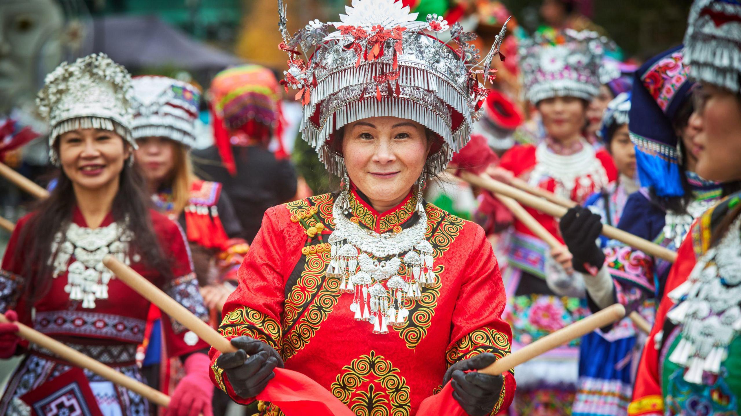 A group of East Asian women in bright embroidered traditional outfits and silver tiered headdresses holding woodens drumsticks