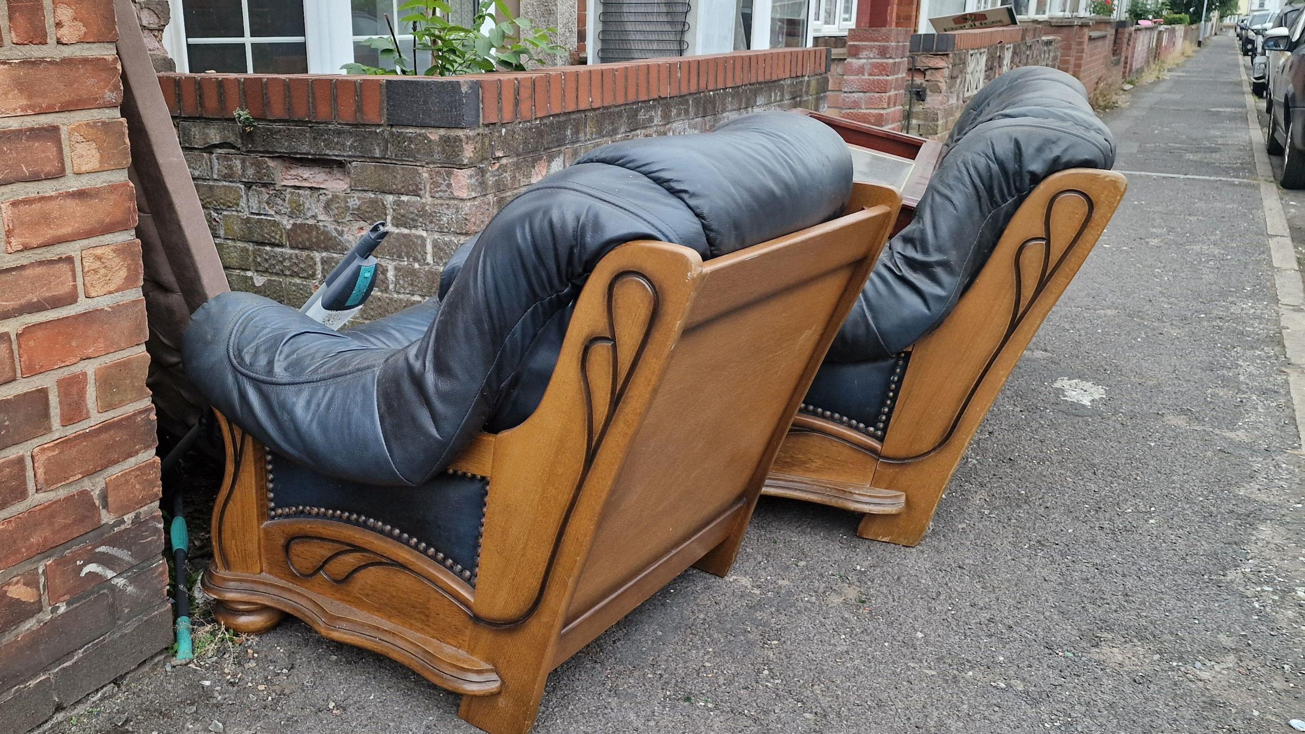 Two dark coloured and wooden armchairs block a pavement on a residential street.