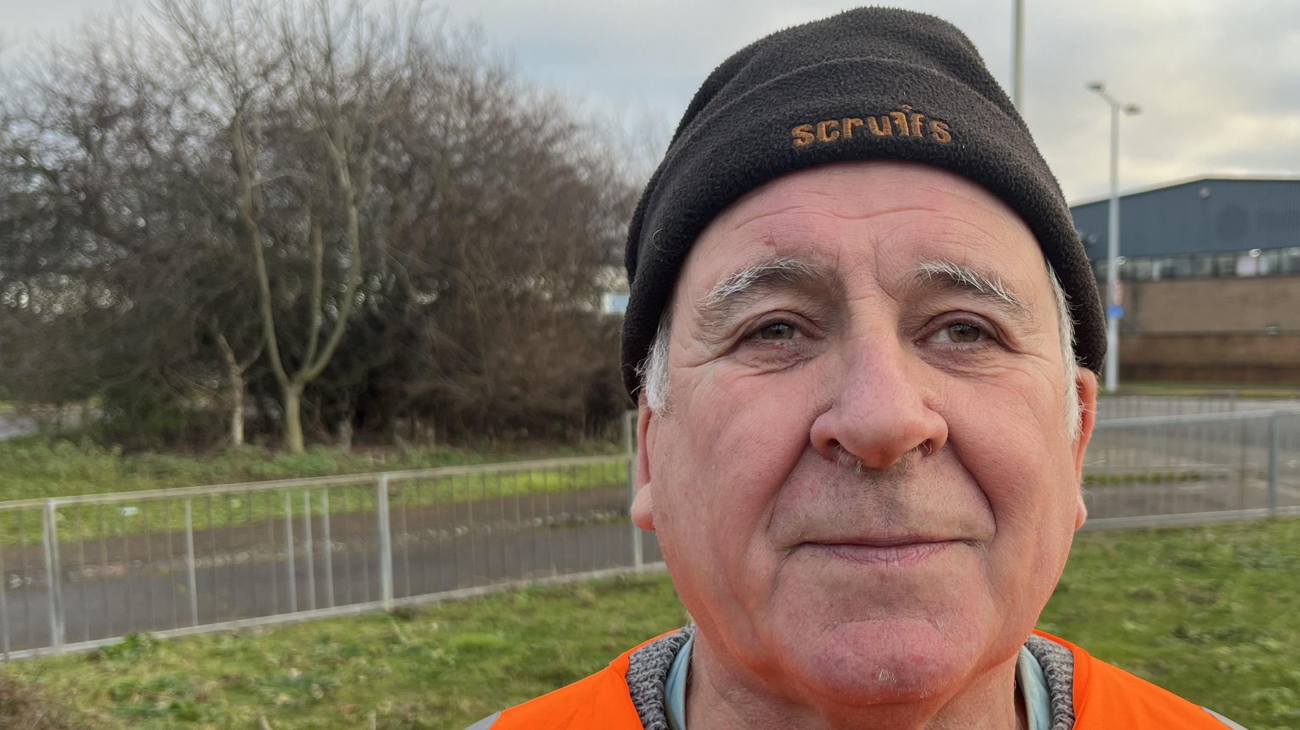 Mark Fishpool, 68, has a black beany hat on with some grey hair and side burns visible. He is wearing a hi-viz orange jacket with a grey jumper underneath. An urban street can be seen behind him.