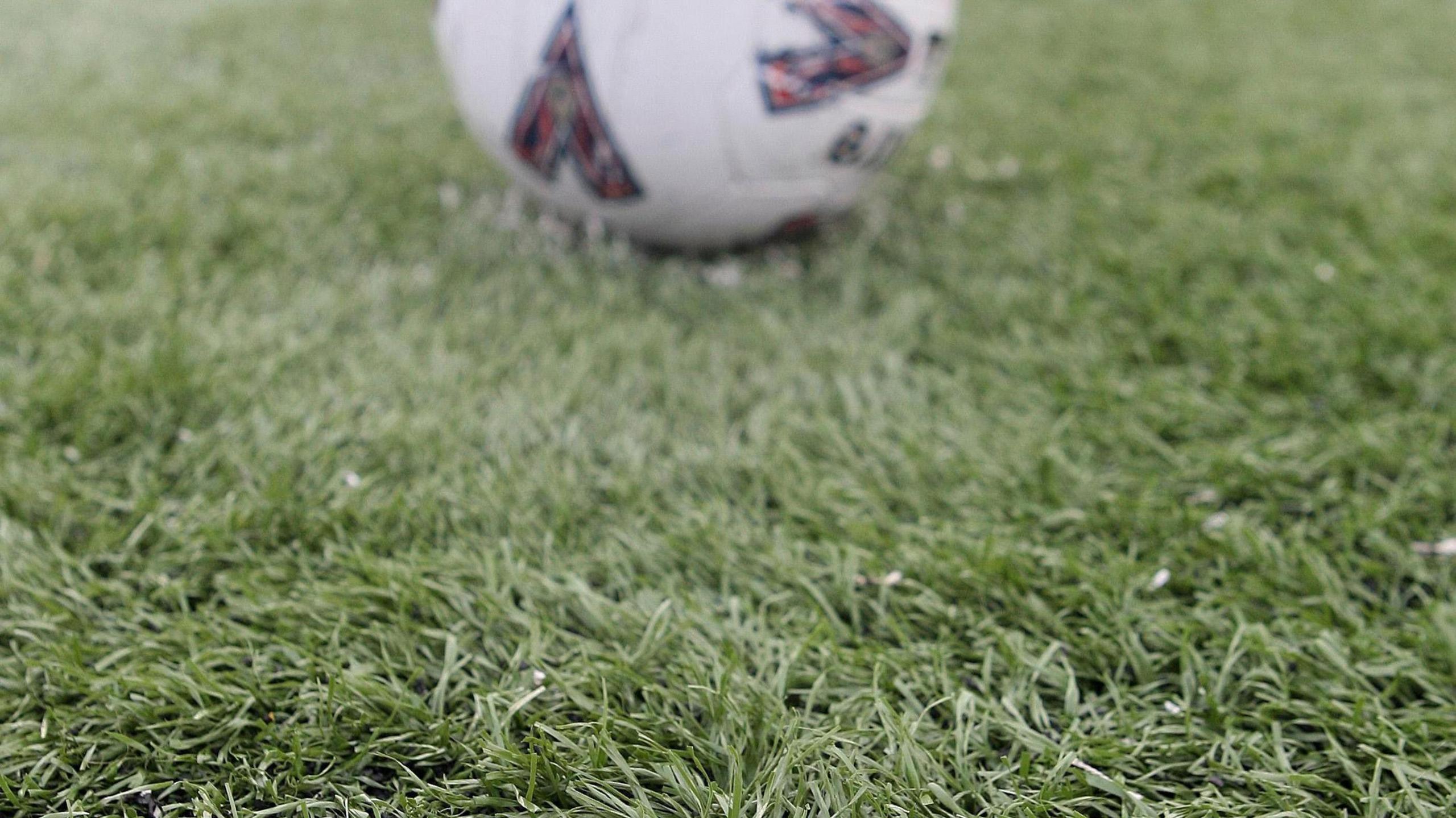 A close-up shot of a football resting on an artificial grass pitch. The pitch is green and focused on it with the white football out of focus in the distance.