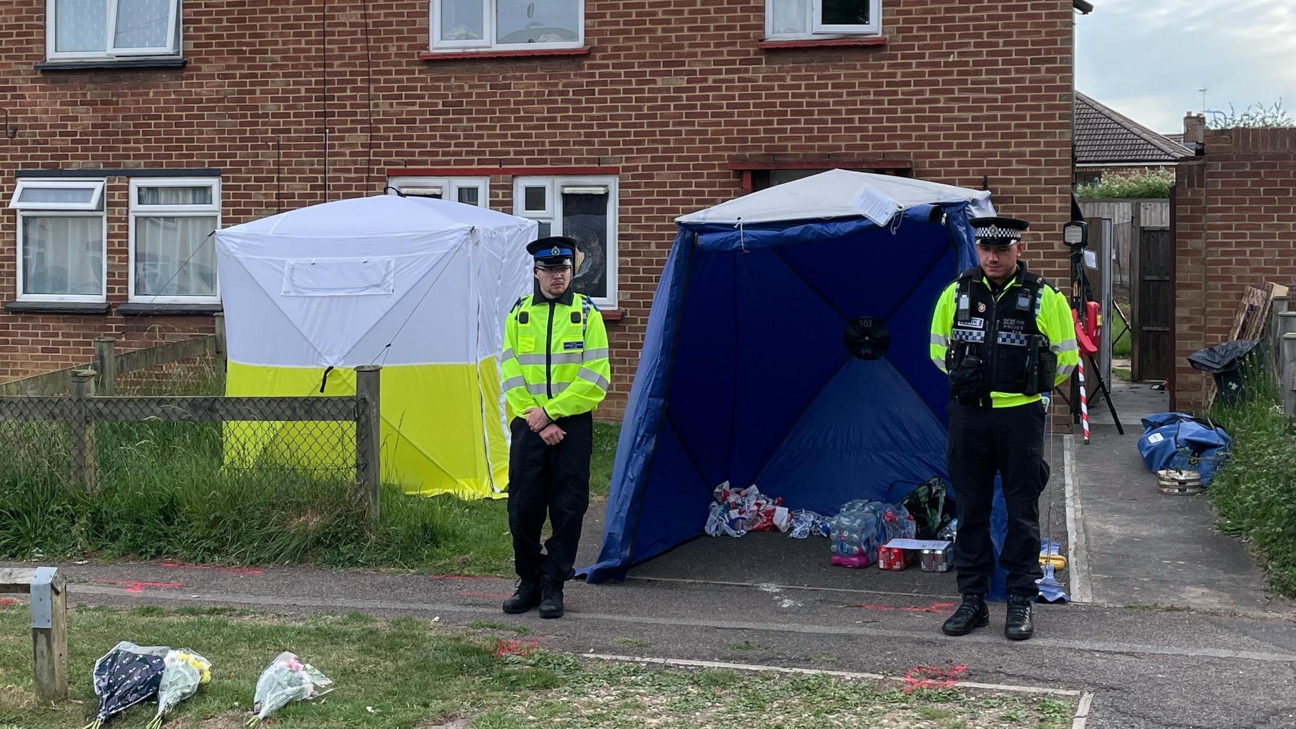 Two police officers and a police tent outside a house. There are flowers laid on the ground and various items are under a blue tent