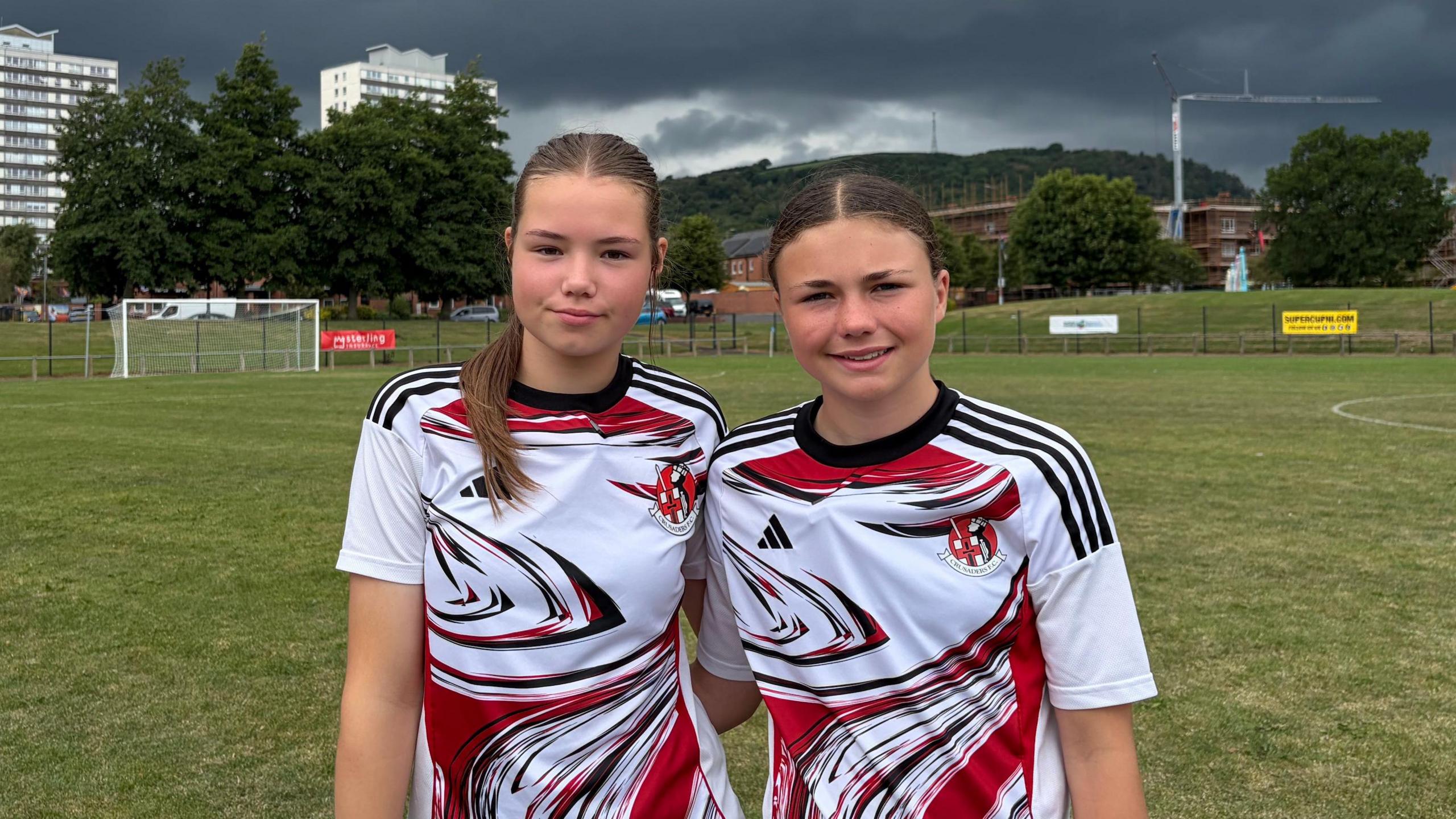 Two girls with brown hair in Crusaders FC away shirts (white with red and black markings) stand on a football pitch. High rise flats can be seen in the background, the sky has dark rainclouds.