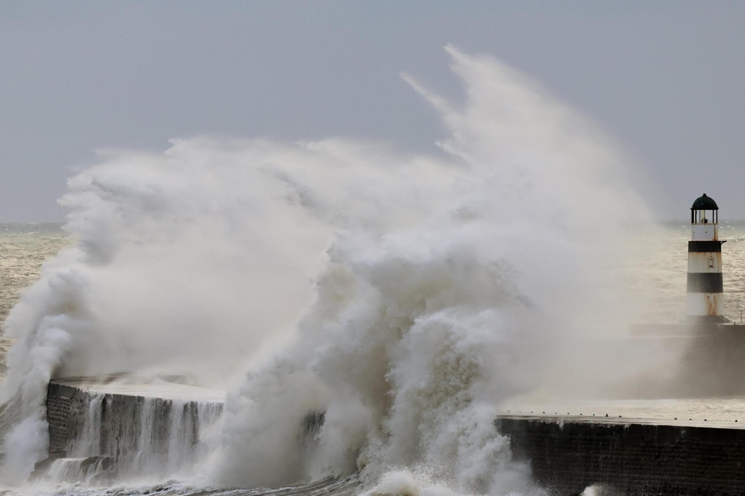 Waves crash up against the pier and its black and white lighthouse.