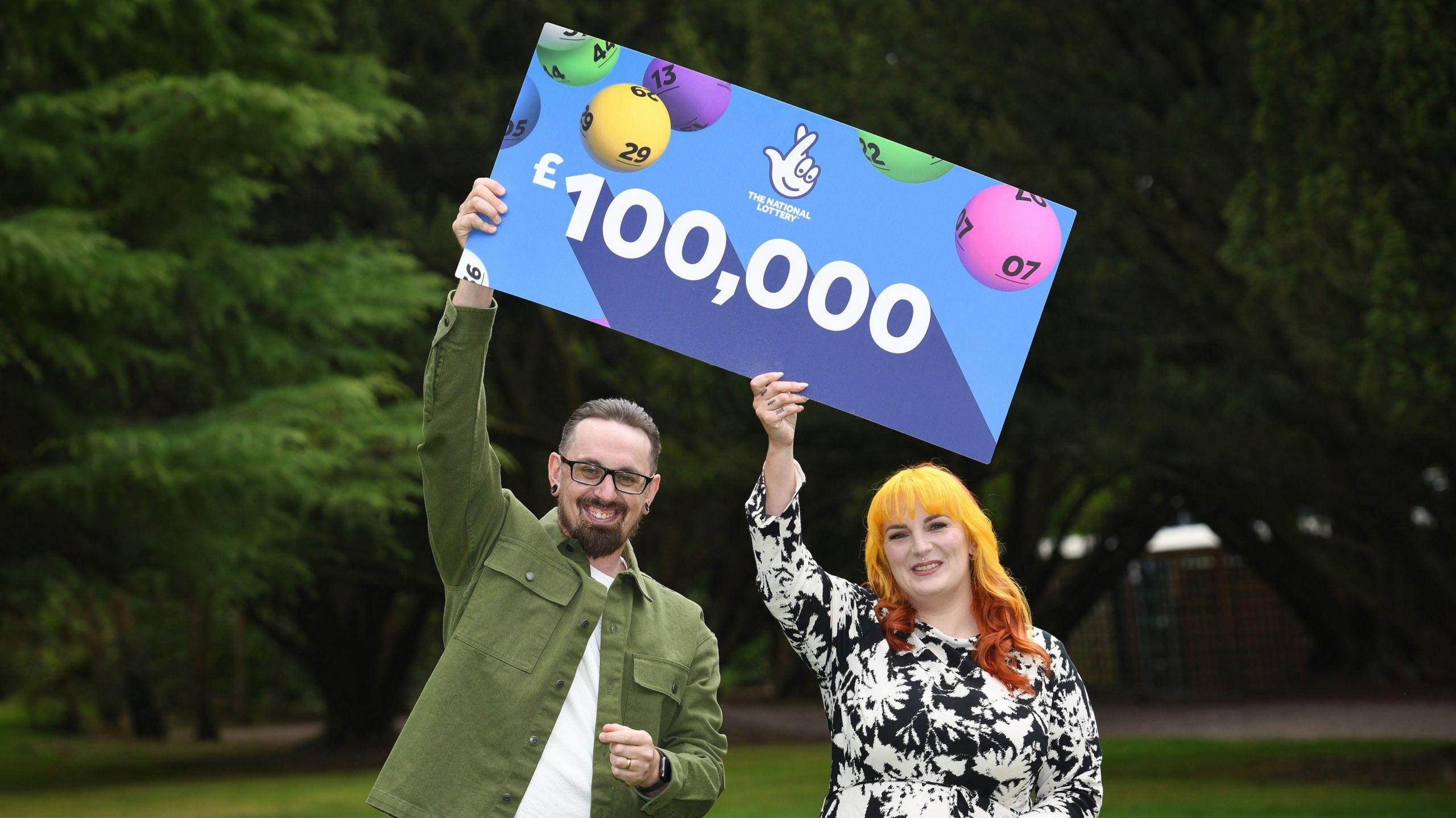 Dan and Kayleigh Cater holding up a sign reading £100,000 with the National Lottery logo on it. There are trees in the background