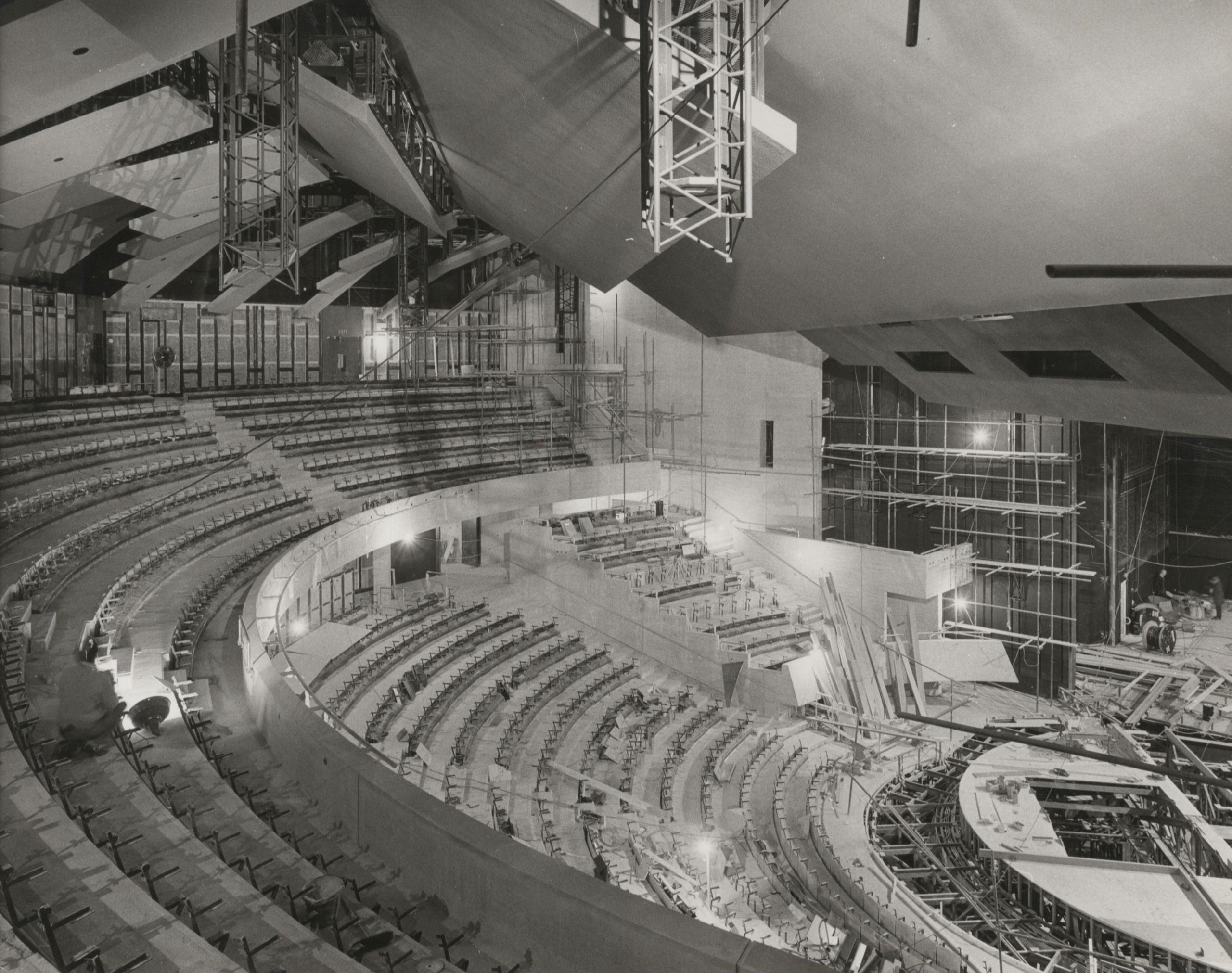 Scaffolding fills a large unfinished interior space inside the National Theatre during construction. There is curved seating in tiers above a stage to the right that is being constructed. 