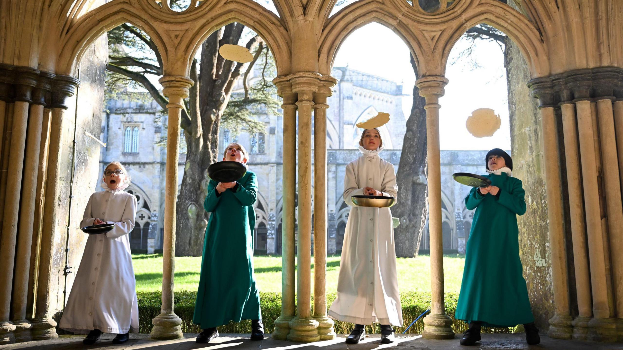 Four young choristers stand in a row under medieval arches with the sun shining through. All have frying pans and are mid-way through flipping pancakes.