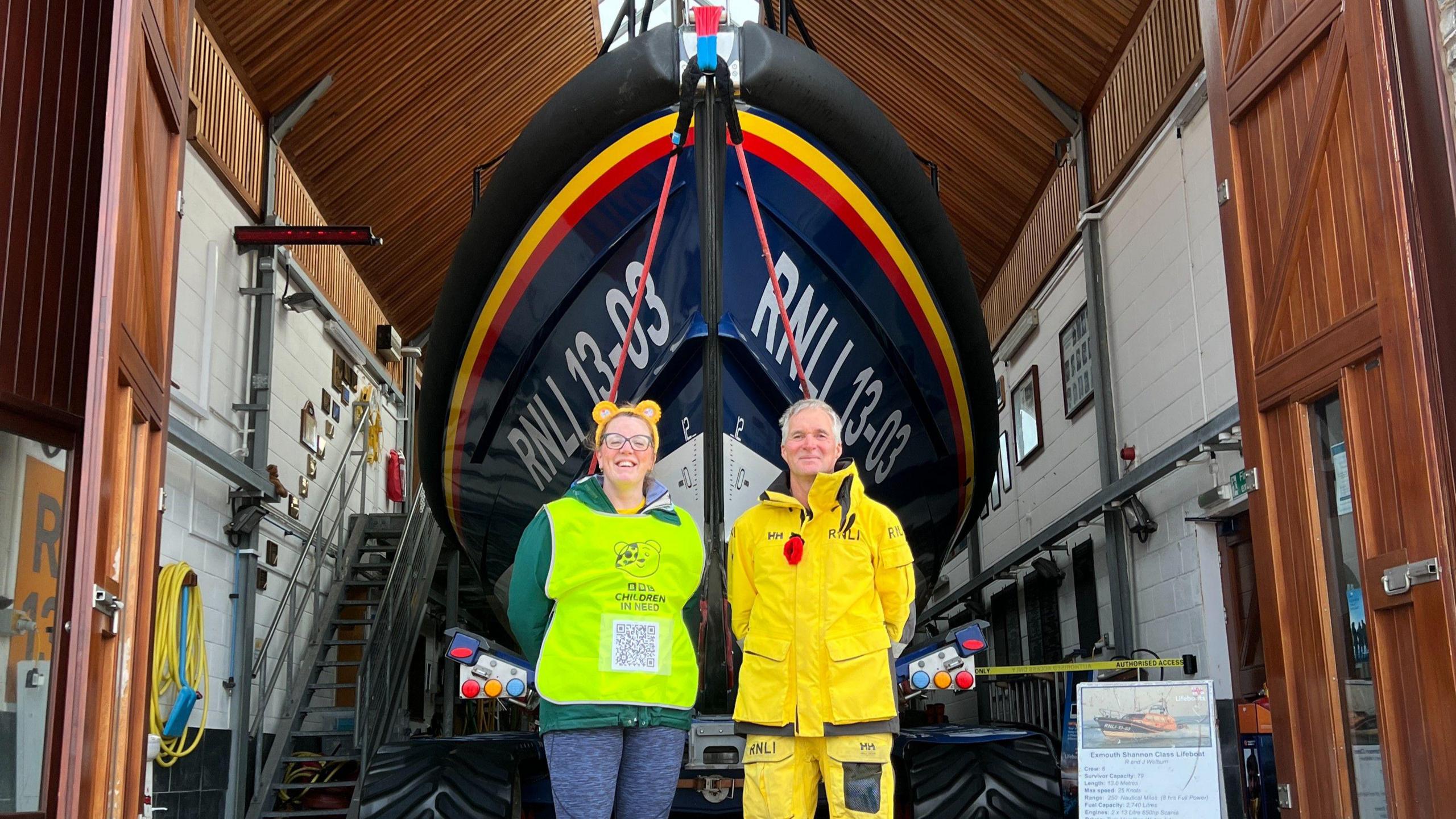 Caroline Densley, who is wearing blue leggings and a high-visibility vest, is standing next to a man in a yellow, all-weather suit. They are standing in front of an RLNI lifeboat.