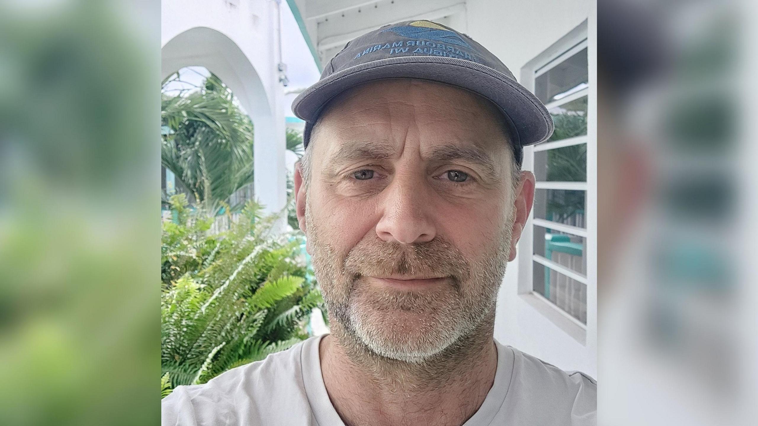 Billy Taylor looks at the camera in a selfie photo. He is wearing a faded blue cap on his head and in the background there are green plant leave and a white building visible.