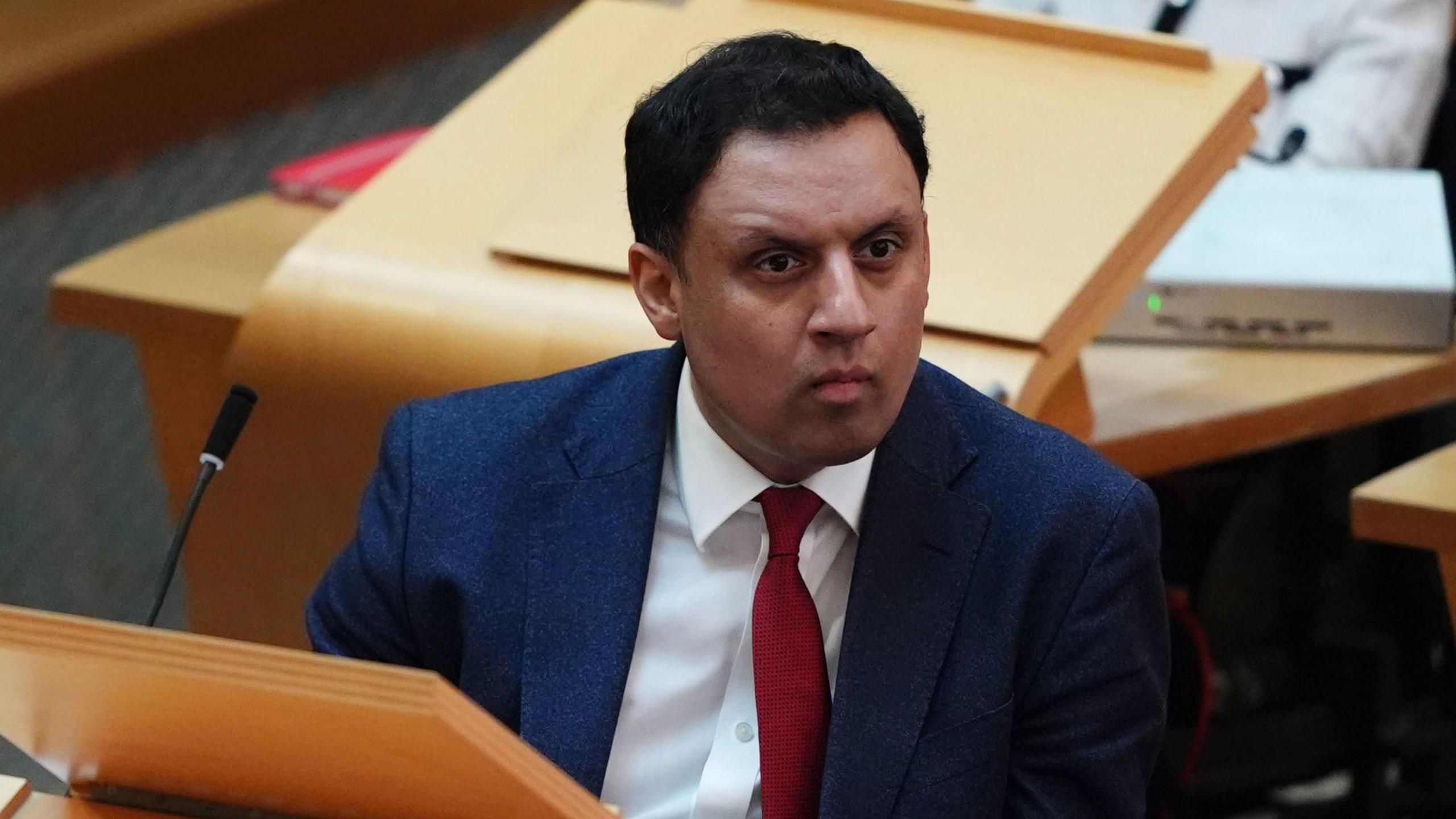 Anas Sarwar has short dark hair and sits in the debating chamber at Holyrood and wears a dark blue suit