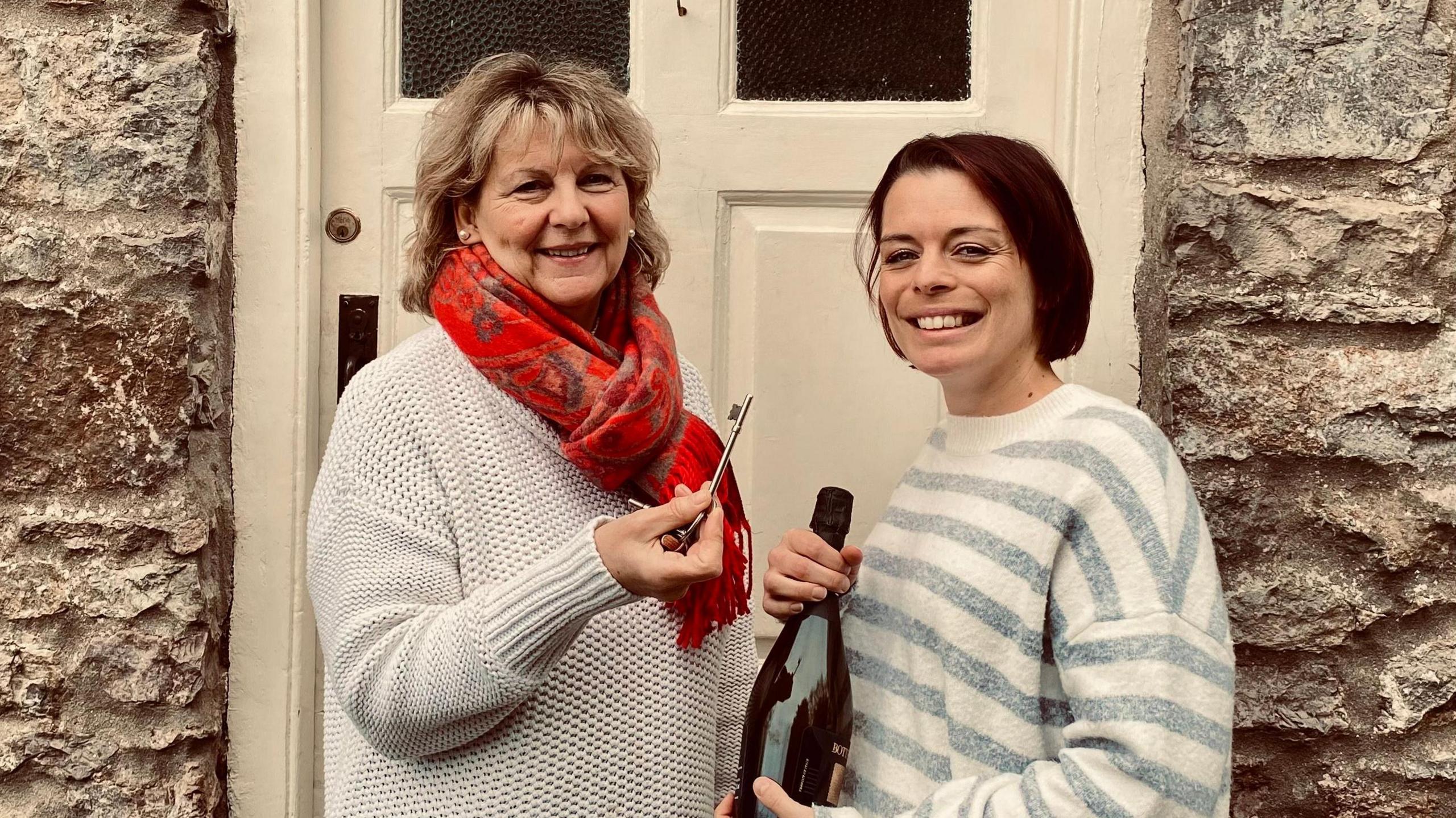 Emma Lomas standing in front of one of the entrances to The Crown Inn with Karen Clements. Emma is holding a bottle of fizz while Karen is handing her a key to the pub. 