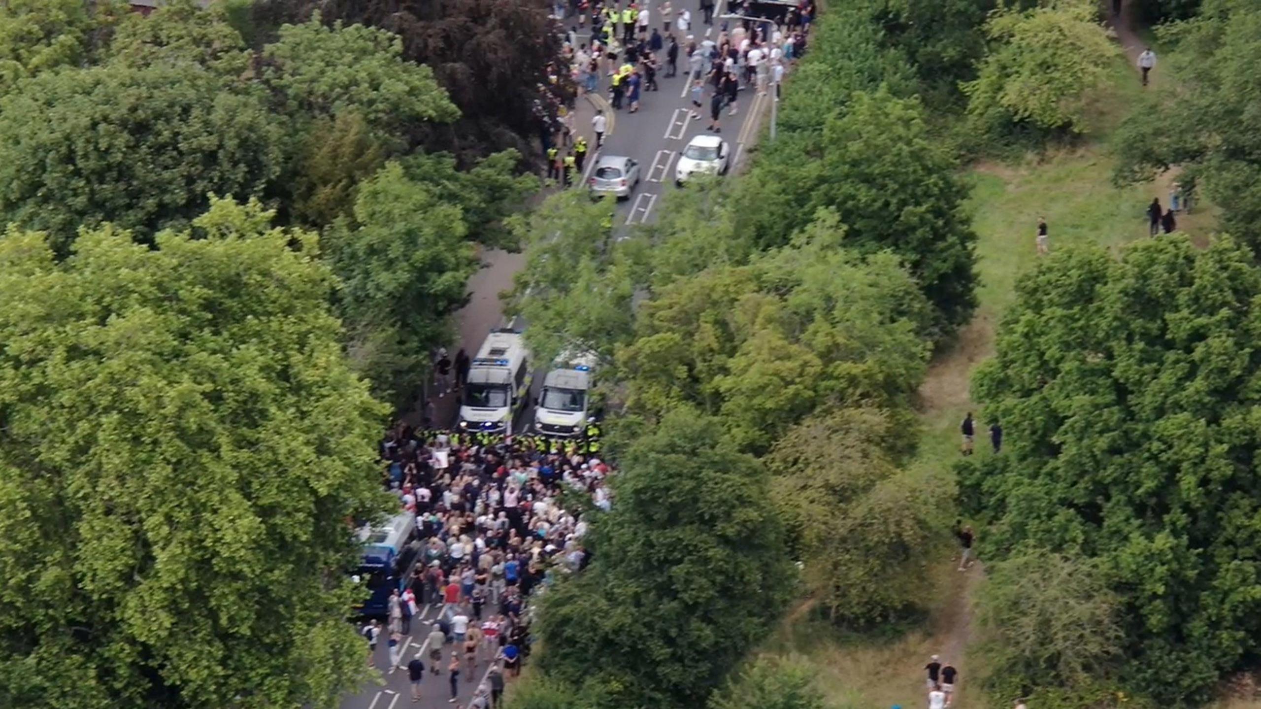 A drone image of two large groups of people being held back by a police cordon. They are standing in a road which is lined by trees and greenery.