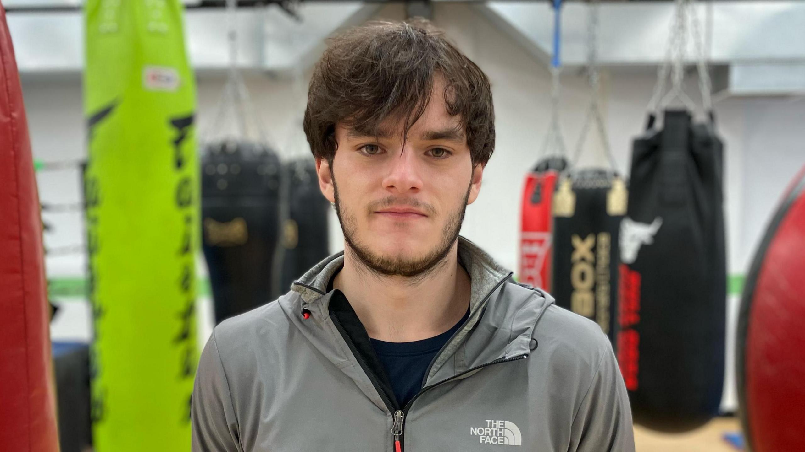 Niall looks into the camera. He has medium-length brown hair down over his forehead and is wearing a grey North Face zipped-up jacket. In the blurred background we can see he is in a boxing gym.