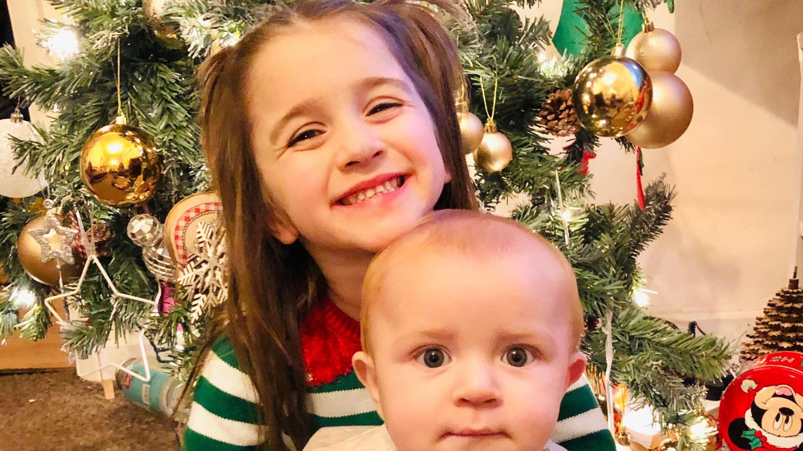 Sophie's five year old daughter Elsie sits in front of a lit up Christmas tree, grinning at the camera. She wears a green and white striped long sleeve top and has brown hair pulled into pigtails. Sophie's one-year-old son Ronnie sits on her lap, he has blue eyes and looks into the camera, sucking his bottom lip.
