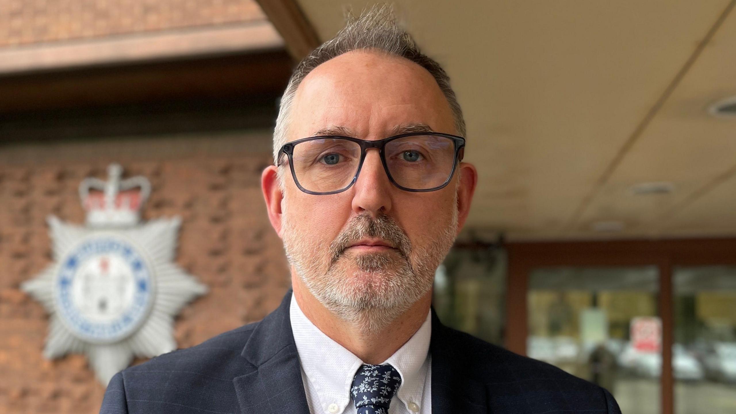 Det Supt Mike Brown looks at the camera. He is standing outside the Suffolk Police headquarters. He has short dark and grey hair and is wearing black glasses. He is wearing a navy suit, white shirt and navy tie. The entrance to the building, which is blurred, can be seen behind him. Behind his left shoulder there is a large police shield on the wall.