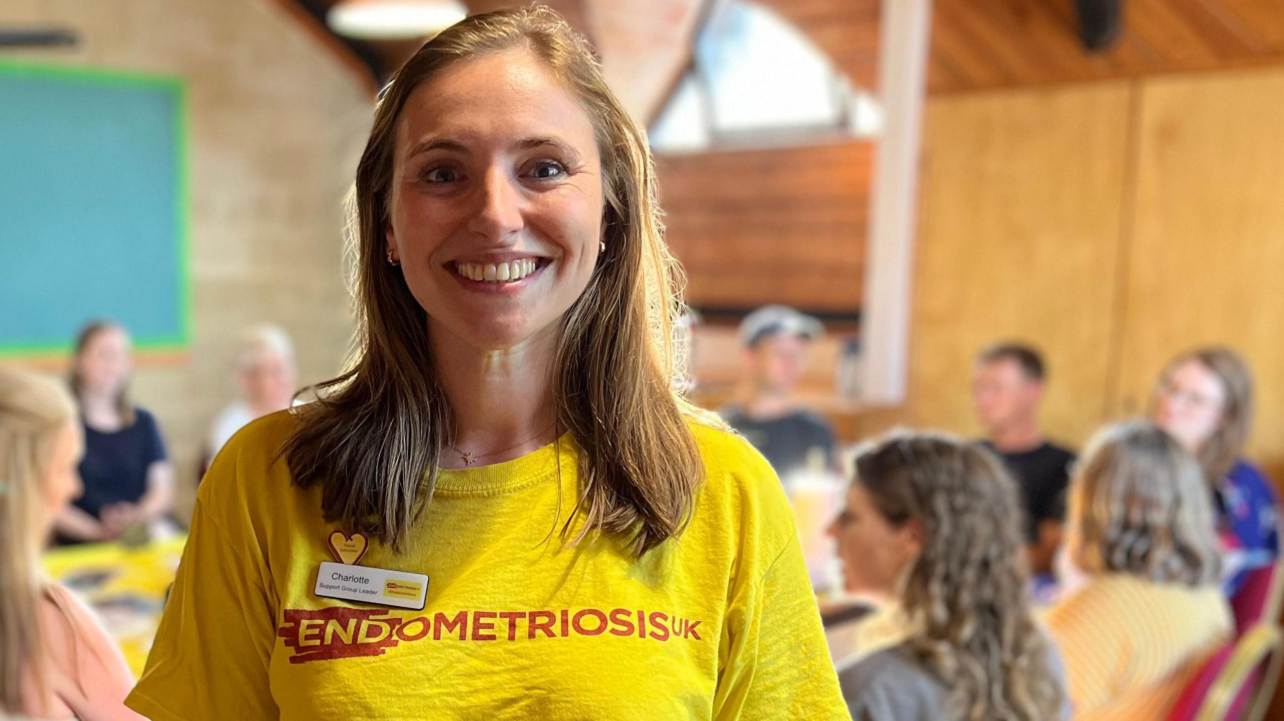 Charlotte smiles at the camera, wearing a bright yellow top with the words "Endometriosis UK" on it in red writing. She has shoulder-length, light brown hair, and is standing in front of a table with mostly women sitting around it talking. 