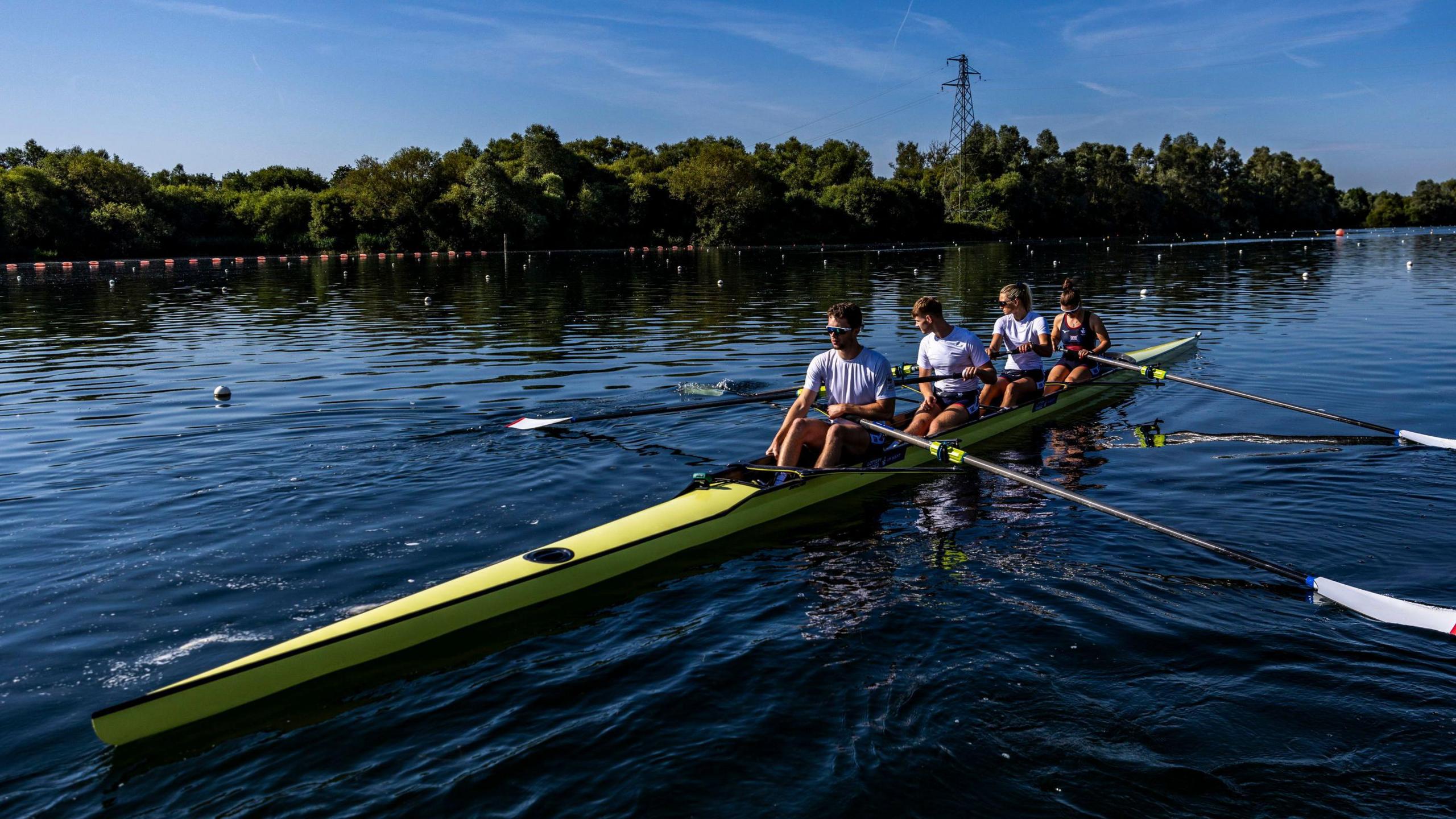 Four athletes dressed in sports gear and all wearing sunglasses sit in a yellow rowing boat on a body of water at ease, each in control of one oar.  