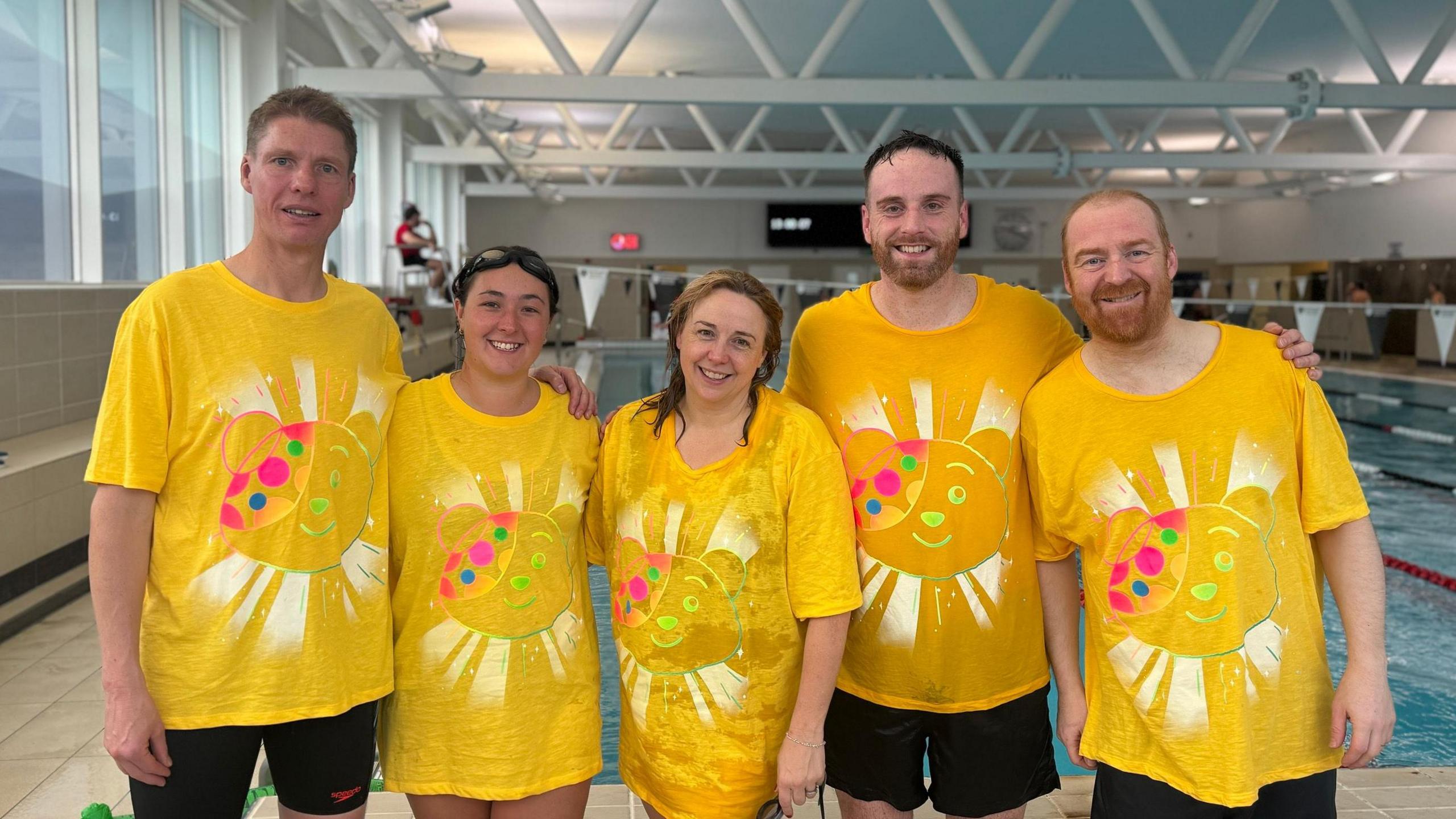 A line of people with wet hair standing in front of an indoor pool. They are wearing yellow T-shirts with Pudsey Bear's face on it. 