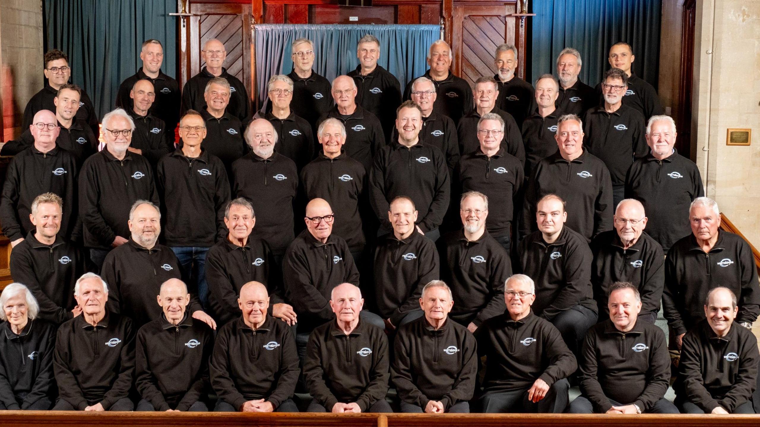 A group photo of a male choir. The members are wearing black shirts. They are sitting in rows and smiling at the camera.