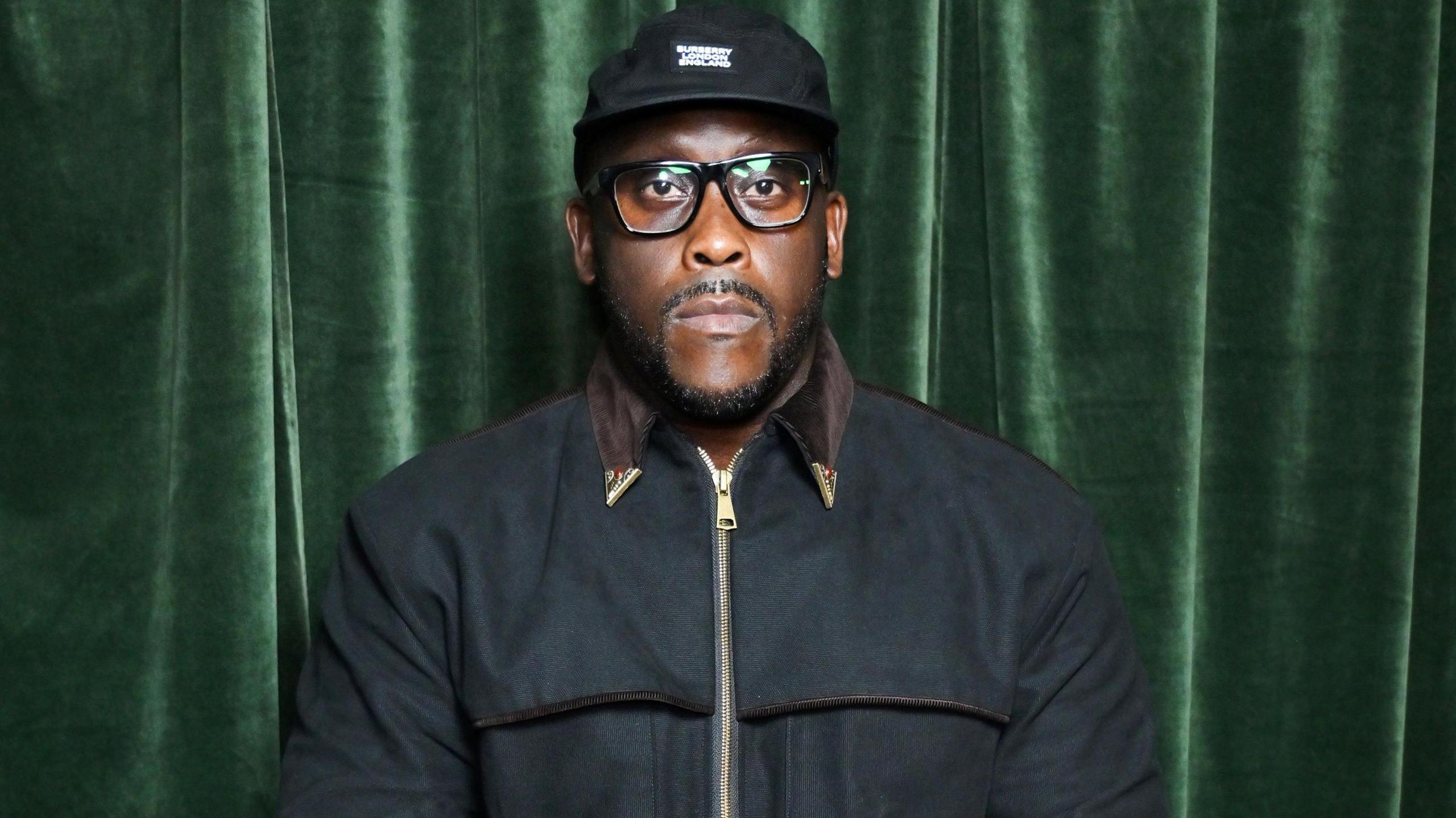 Kenny Allstar pictured in December 2024. He wears a black cap with a black zipped-up jacket. He has black rimmed square glasses and a trimmed moustache and beard. He's pictured with a serious expression in front of a green velvet curtain. 