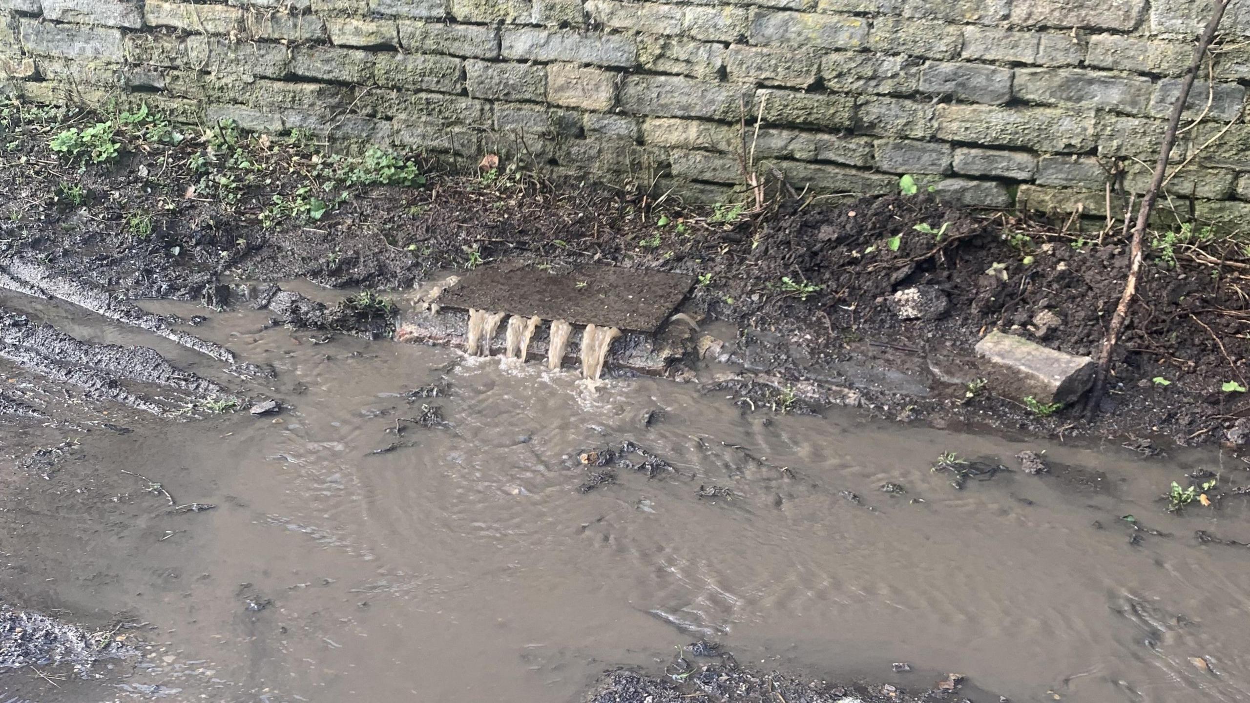 In the centre of the shot is a drain which isn't fully closed,  with brown, discoloured water flowing out on to the road, which is also covered in the same colour water. 