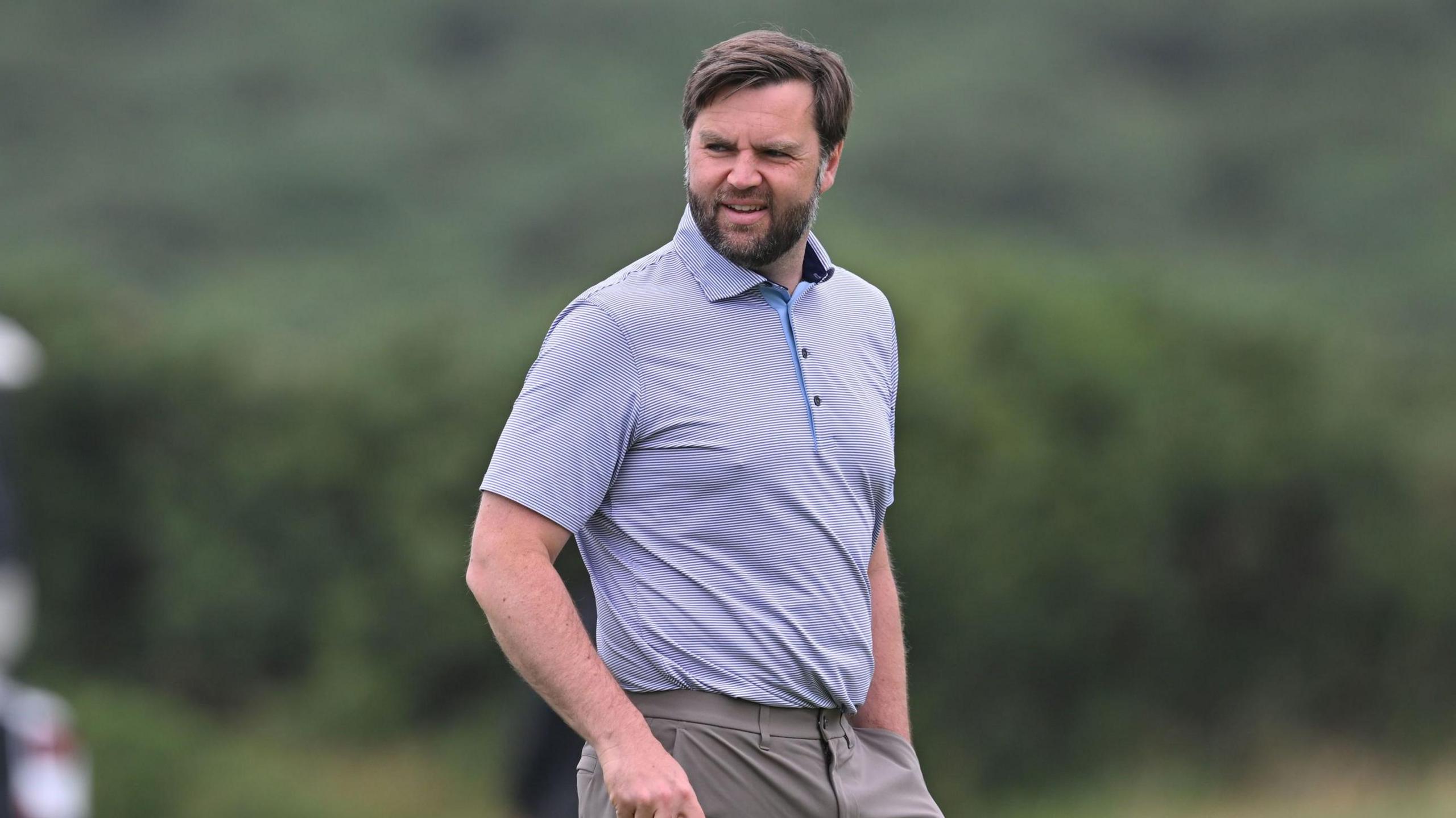 JD Vance looking slightly off camera. He is on a golf course. He is only visible from the waist up. He is wearing a light-coloured polo shirt with a thin horizontal pinstripe design. He is wearing dark beige trousers. He has dark hair and a dark beard. He is holding a golf club in his right hand, which is just out of shot and has his left hand in his pocket.