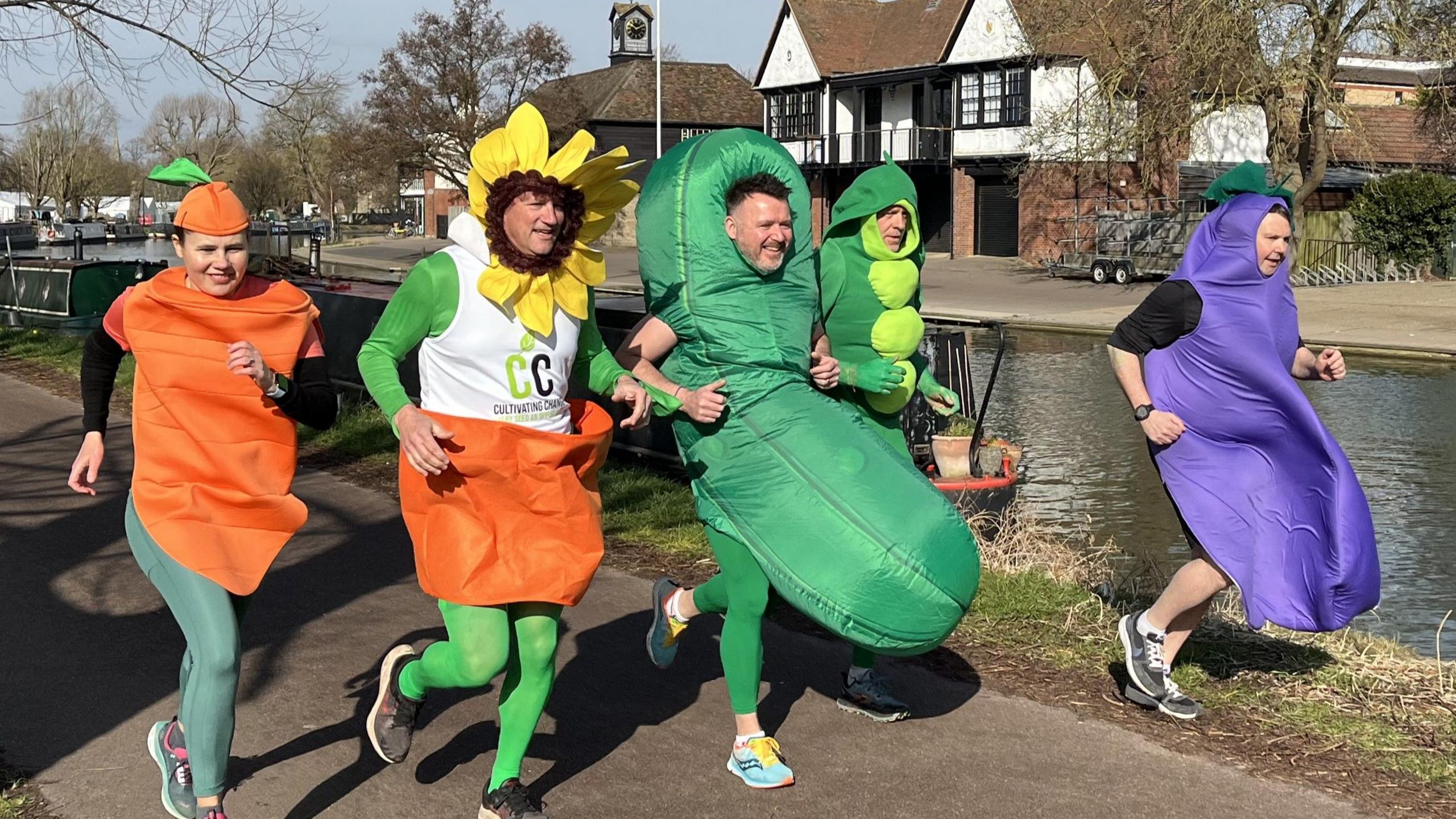 The group dressed as their respective vegetables and the sunflower running along a river. They have smiles on their faces as they run.