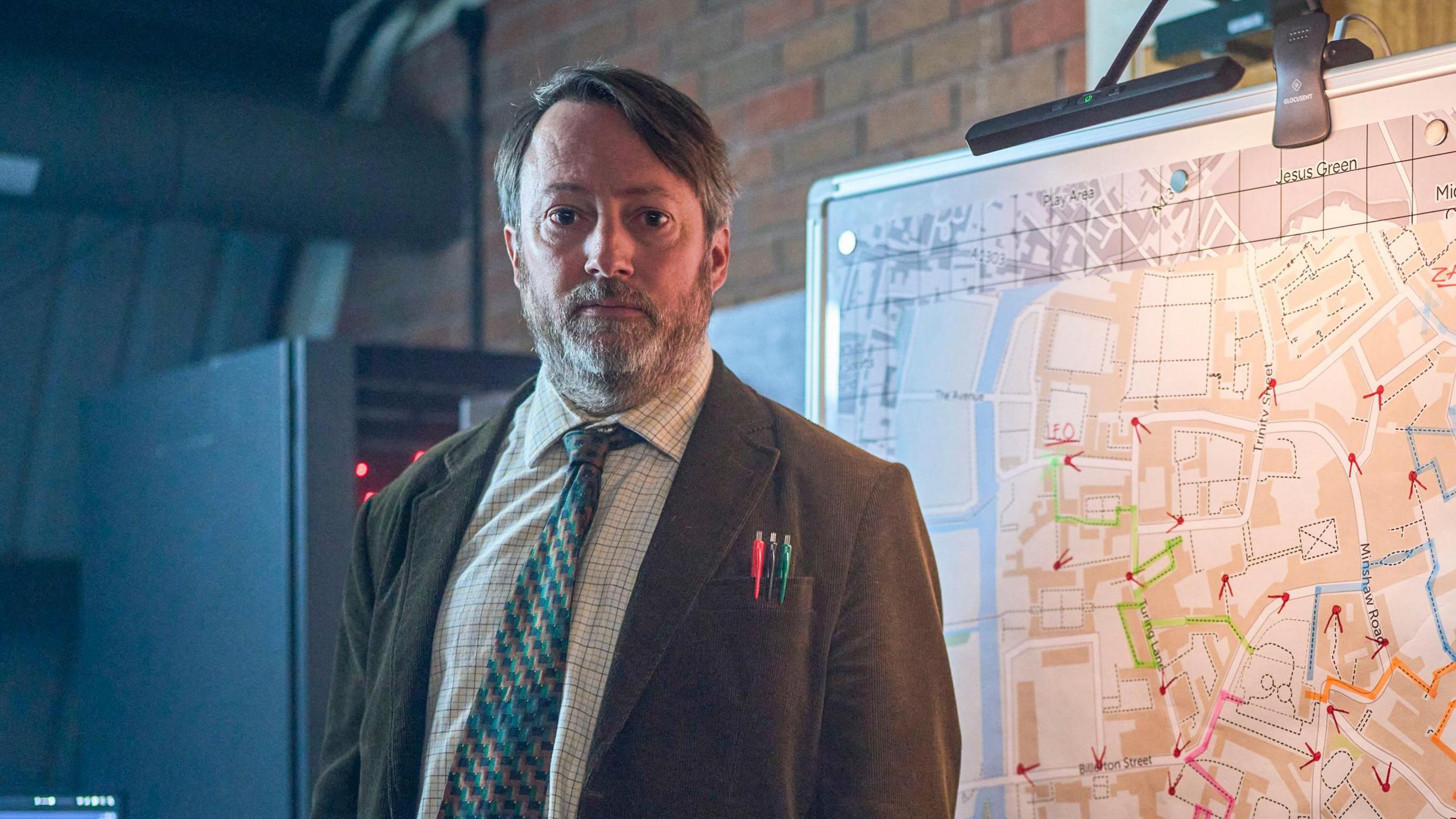 David Mitchell in a brown jacket, shirt and tie looks concerned at the screen