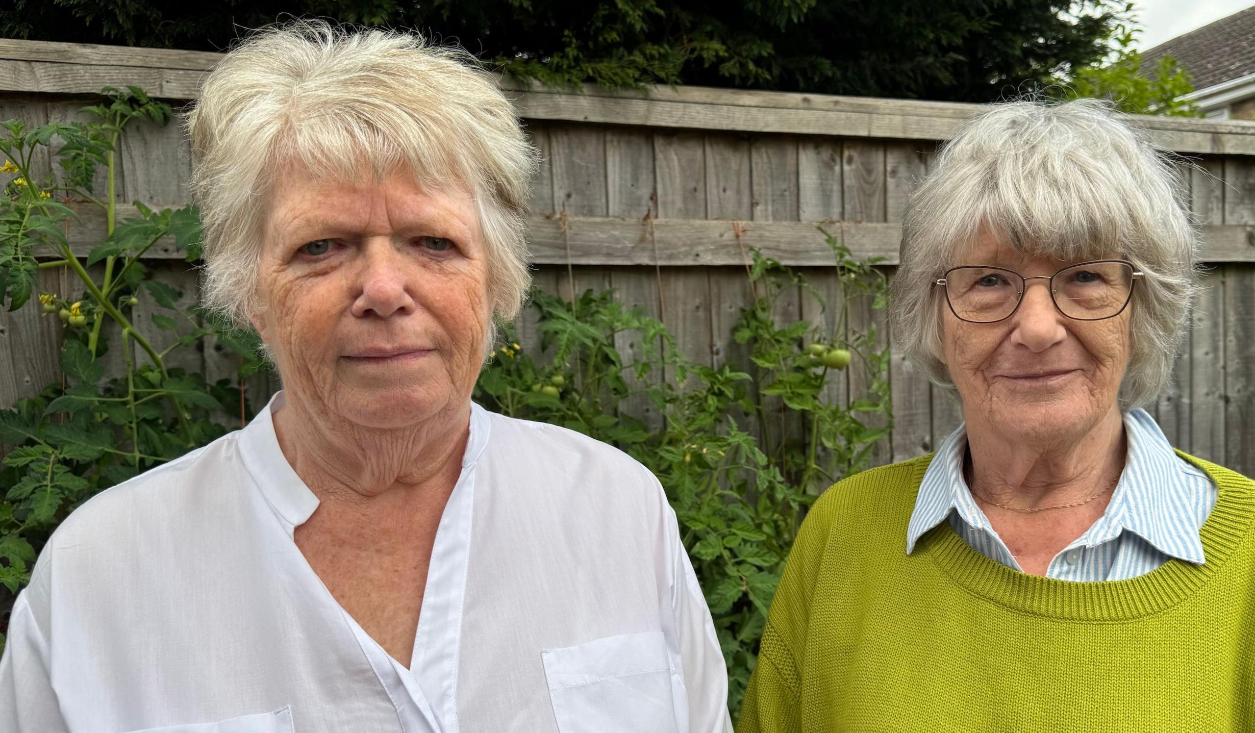 Heather and Gill are women in their 70s. Both have grey hair. Heather is wearing a white shirt, Gill is wearing a blue and white striped shirt and a lime green jumper. They are standing next to each other in a garden and looking at the camera.