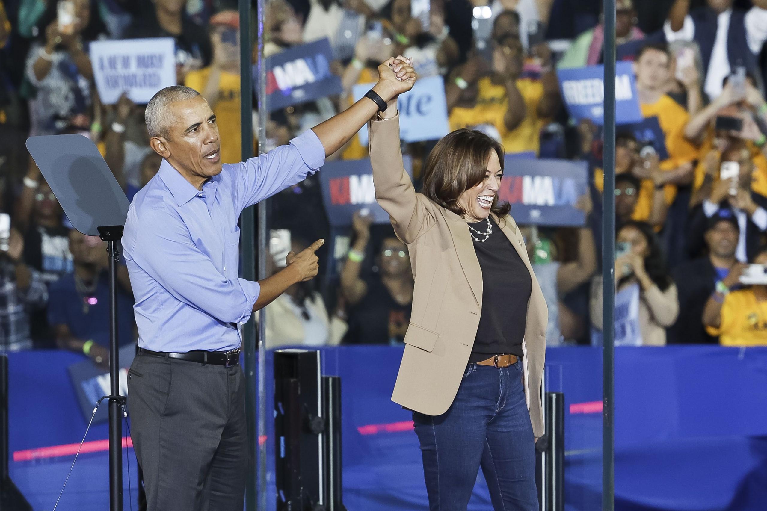 Former US President Barack Obama celebrates by holding up Kamala Harris' hand in the air and gestures towards her on stage at a rally