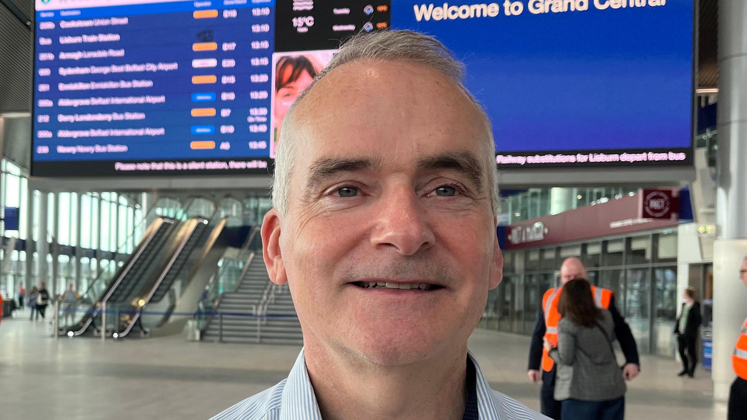 Chris Conway is looking past the camera. He has grey hair and wearing a blue and white shirt. Behind him is a huge blue board with departure times. The foyer, he's standing in, is a vast space with escalators and a closed coffee shop. There are some people in high-vis jackets and some passengers in the back-ground.