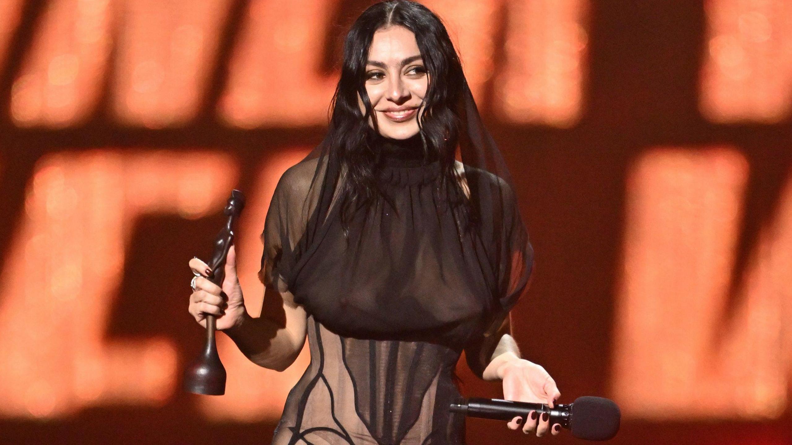 Charli XCX is smiling on stage after receiving a Brit Award statuette, which she is holding in her right hand. She is wearing a see-through black dress and is holding a microphone in her left hand.